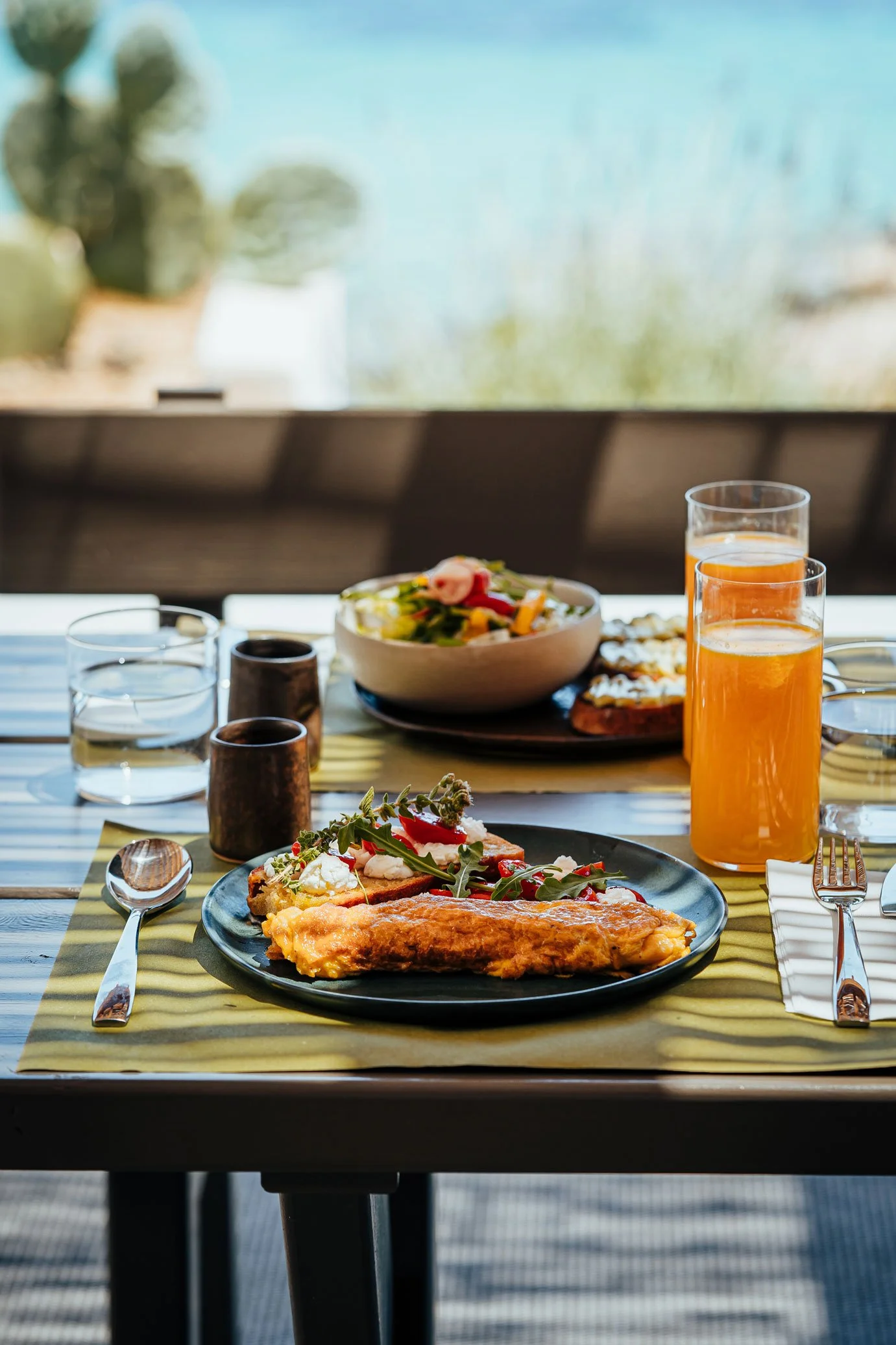 A breakfast table set with a plate of fried fish, toast with strawberries and whipped cream, and a salad, along with two glasses of orange juice, water glasses, cutlery, and salt and pepper shakers, with a window in the background showing an outdoor 