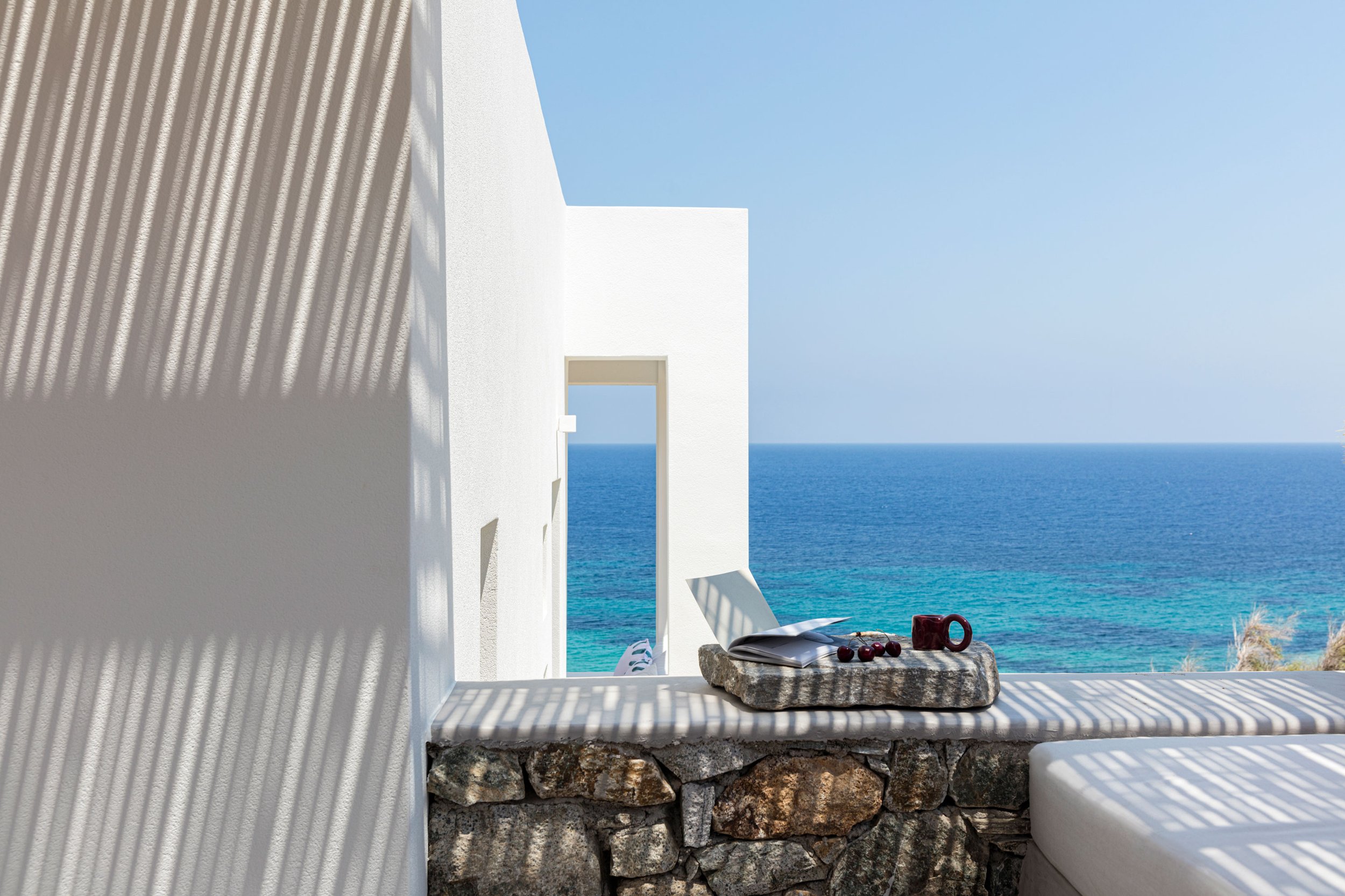 A white building with a terrace overlooking the ocean. Shadows cast by a pergola create striped patterns on the wall and surface. A stone ledge holds a tray with a magazine, cherries, and a red mug, with the blue sea and sky in the background.