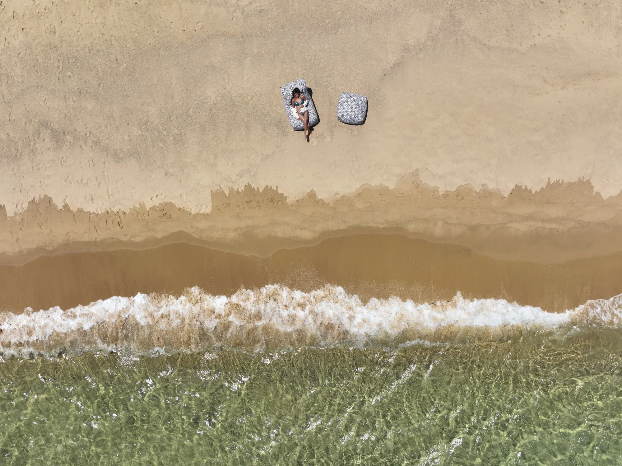 An aerial view of a person lying on a beach blanket with a child, near the shoreline with waves and greenish water.