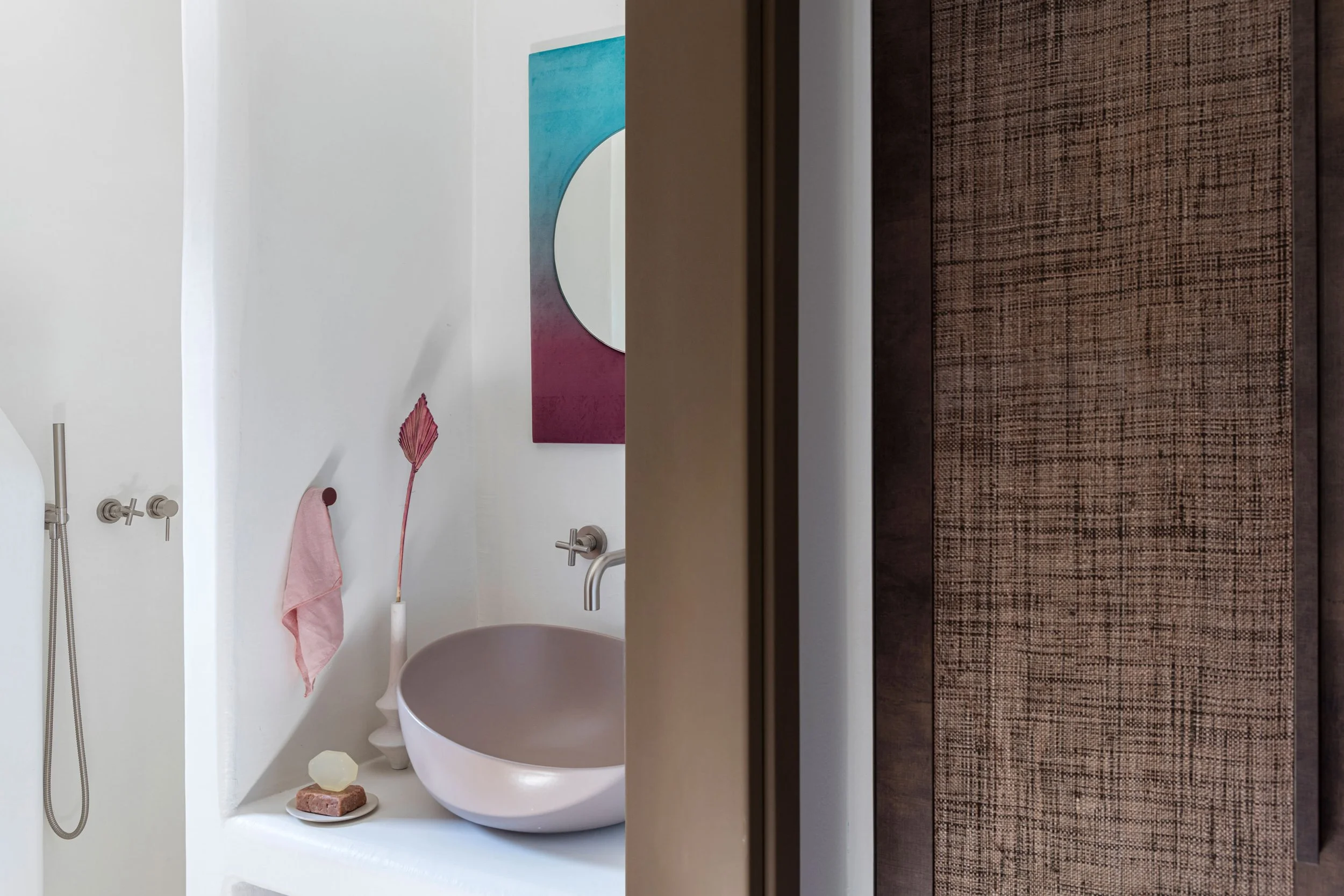 A bathroom with a pink vessel sink, a round mirror, a painting with blue, white, and pink colors, a pink towel, a white vase with a pink plant, soap on a dish, and brown textured wall paneling.