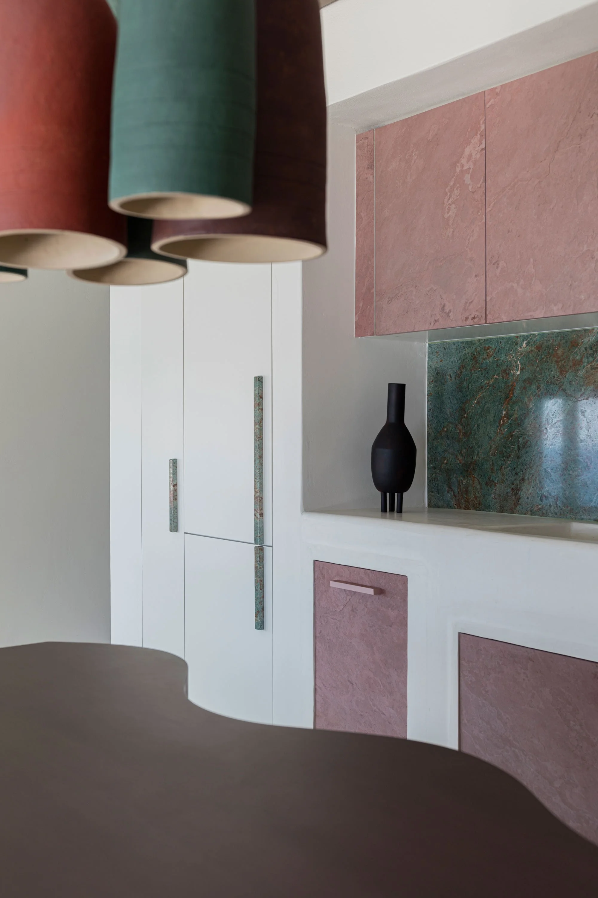Close-up of a modern kitchen with pink and green marble countertops, white cabinets with textured handles, and a black decorative vase on the counter. Overhead, there are several hanging pendant lights in different shades of red, green, and blue.