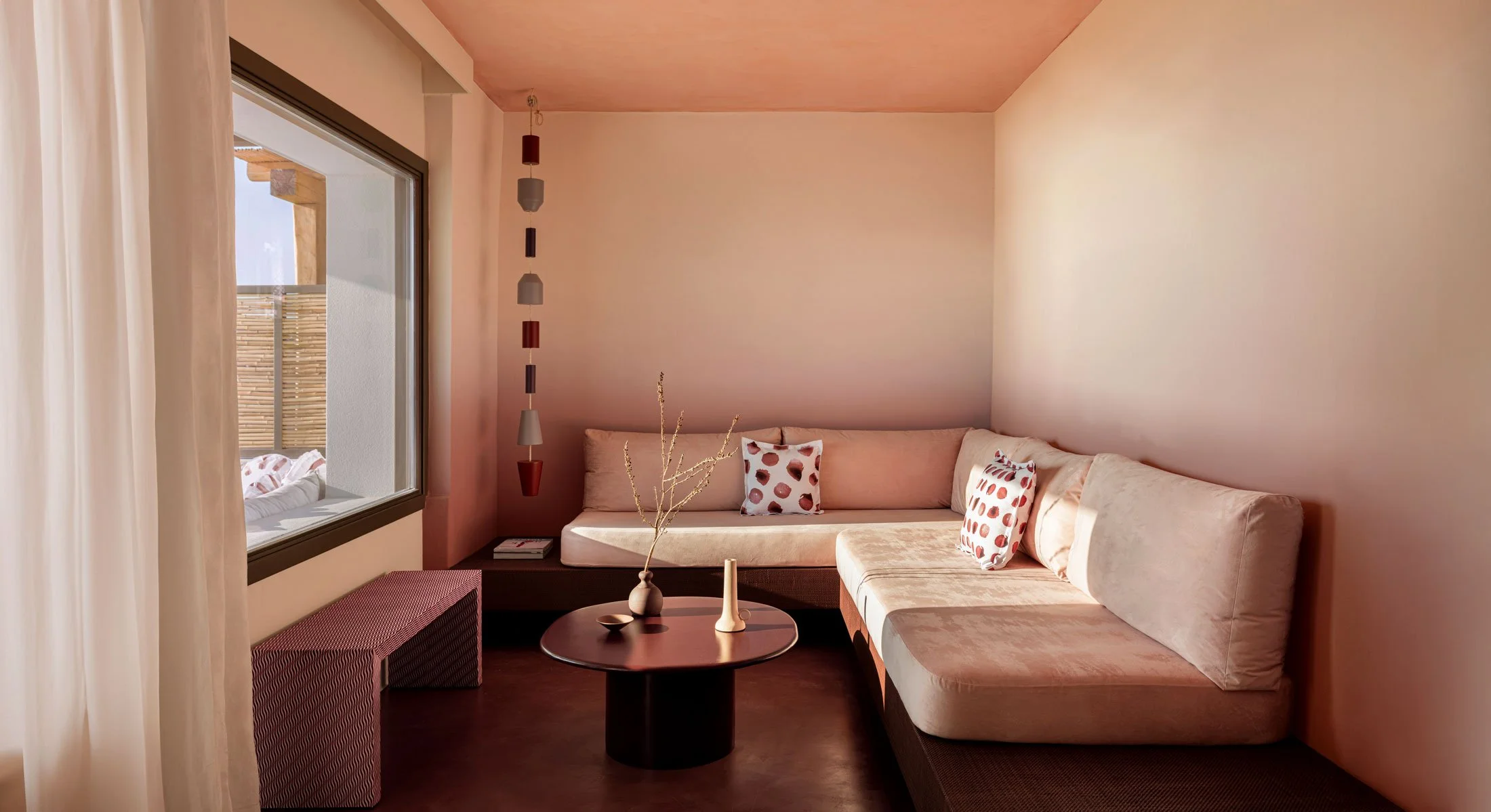 Cozy living room with beige sectional sofa, polka dot pillows, round black coffee table with vases, and hanging pendant lights, next to large window with beige curtains.