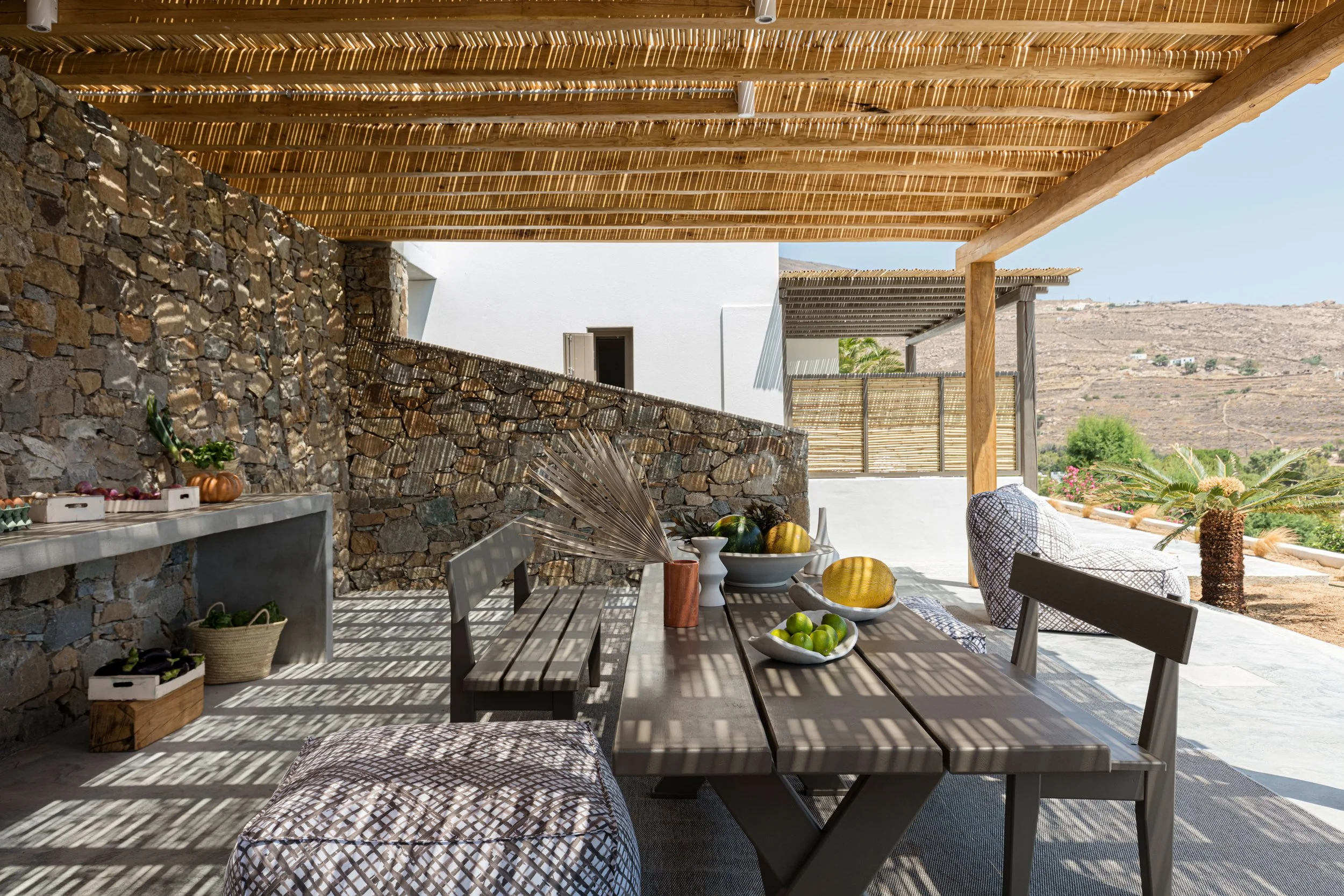Outdoor patio with a wooden dining table, benches, and cushioned chairs, shaded by a bamboo pergola, overlooking a desert landscape.