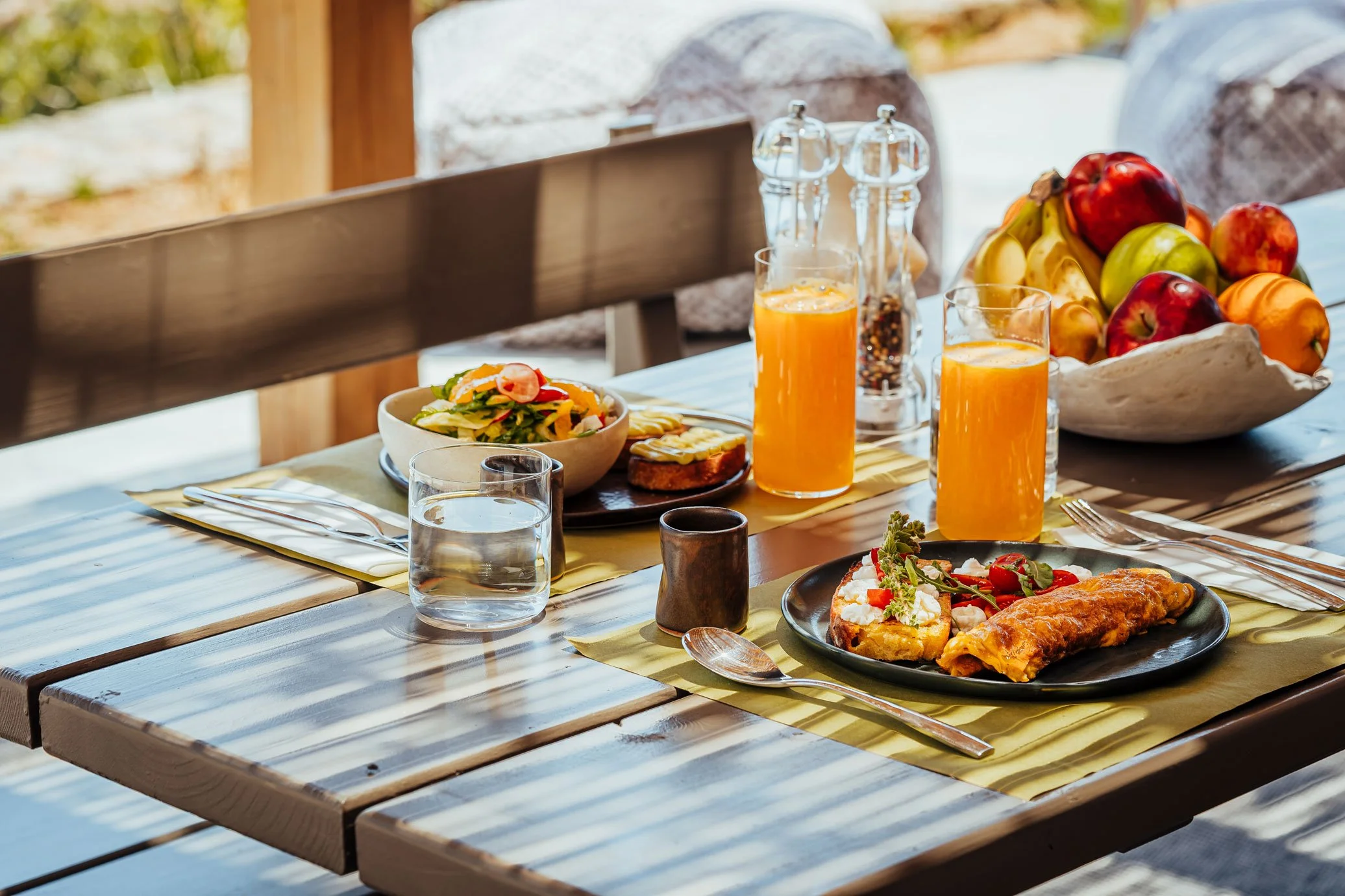 A wooden outdoor table set with breakfast, including plates of eggs and vegetables, glasses of orange juice and water, a bowl of assorted fresh fruits, salt and pepper shakers, and cutlery, with sunlight filtering through nearby trees.