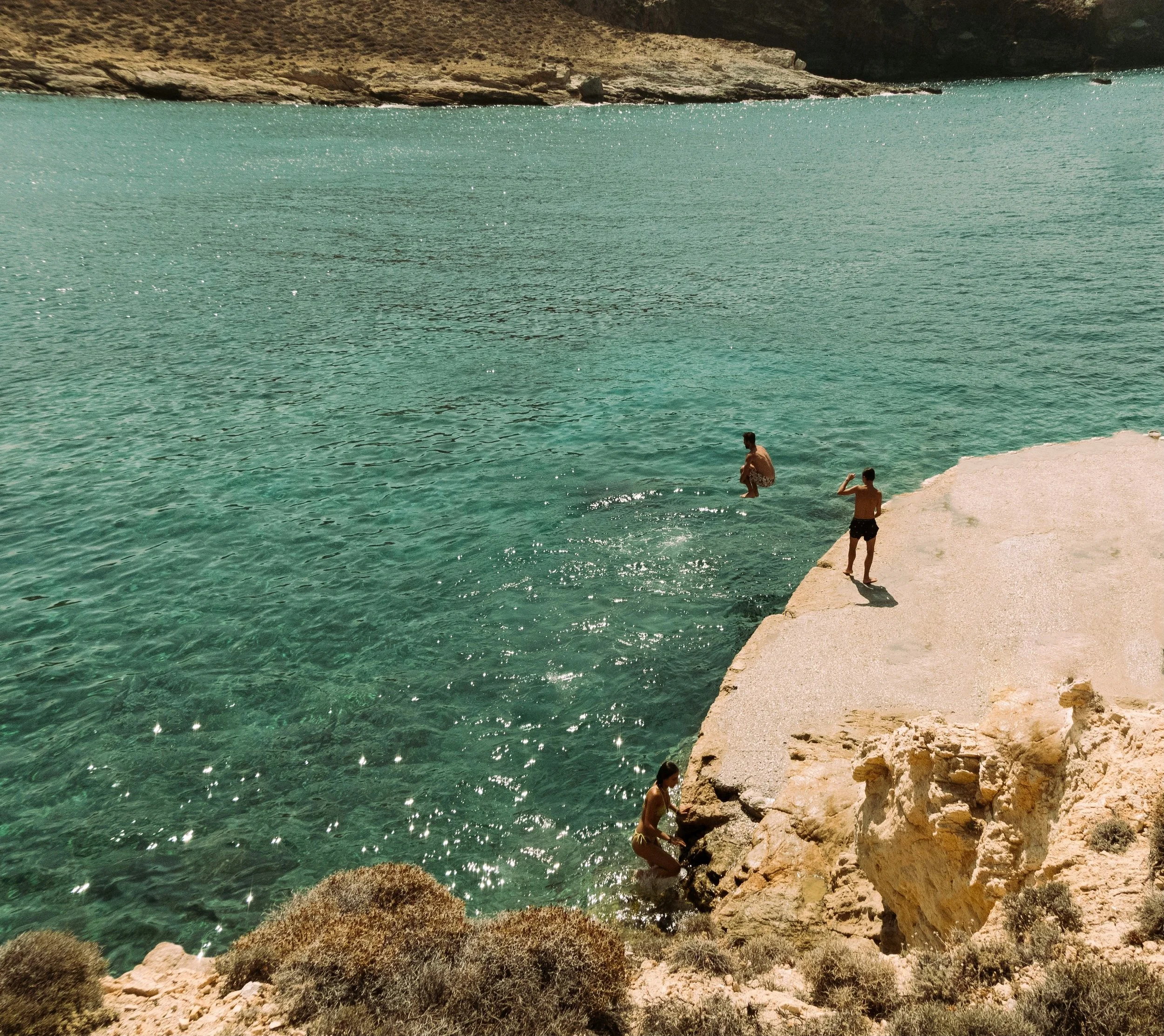 People swimming and wading in a turquoise body of water near a rocky shoreline, with two children standing on a concrete platform over the water and one person sitting on rocks near the shore, surrounded by desert-like vegetation.
