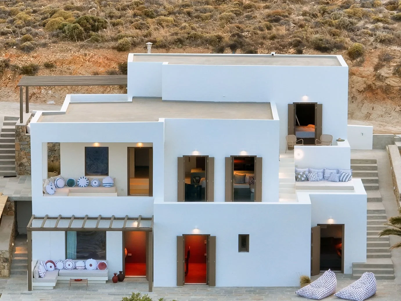 Modern white multi-level house with multiple outdoor seating areas, open windows with shutters, and steps leading to different levels, set in a desert landscape.
