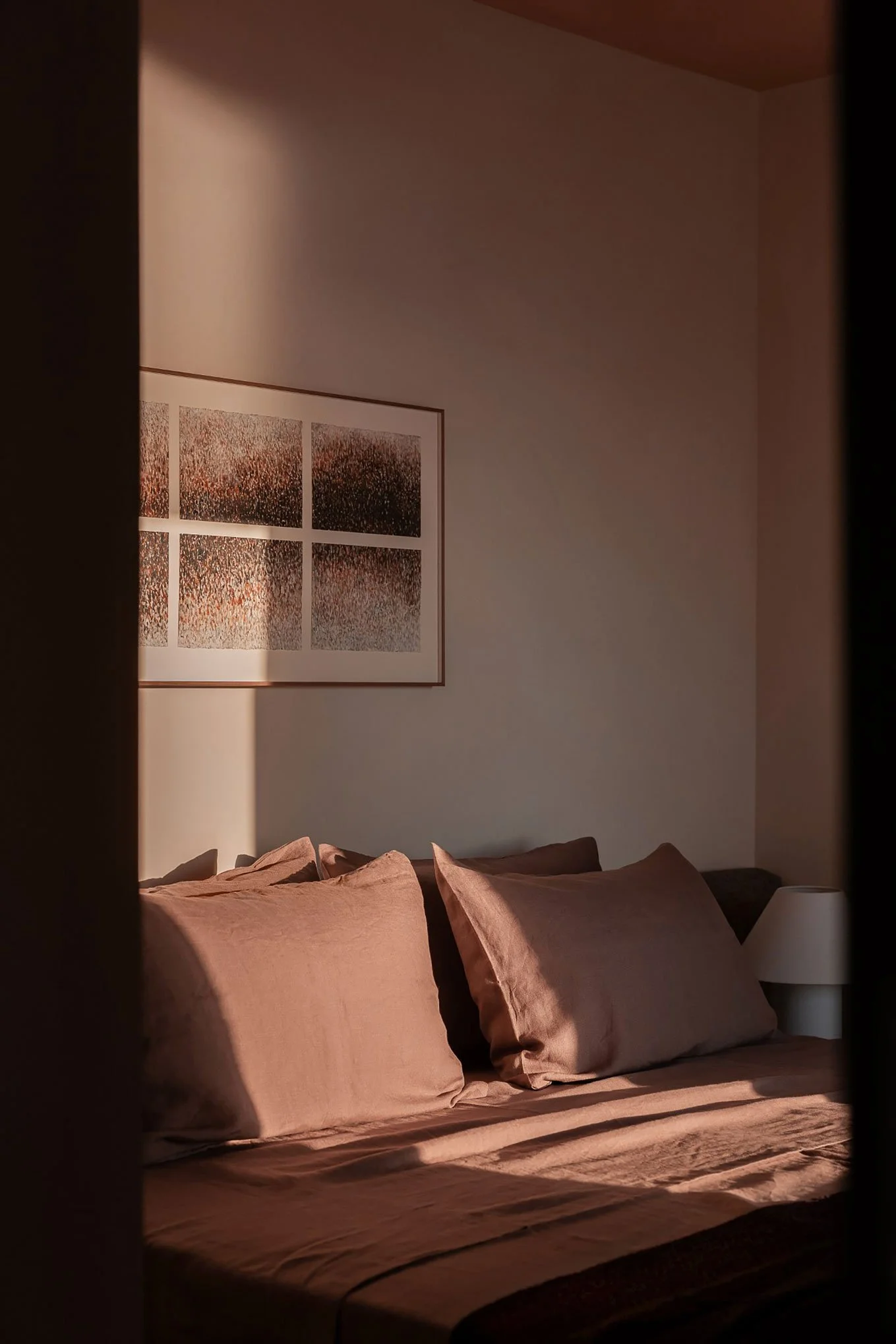 A neatly made bed with beige pillows and a beige bedsheet, seen through a partially opened door. The room has sunlight casting shadows, a framed abstract artwork on the wall, and a small white bedside lamp.