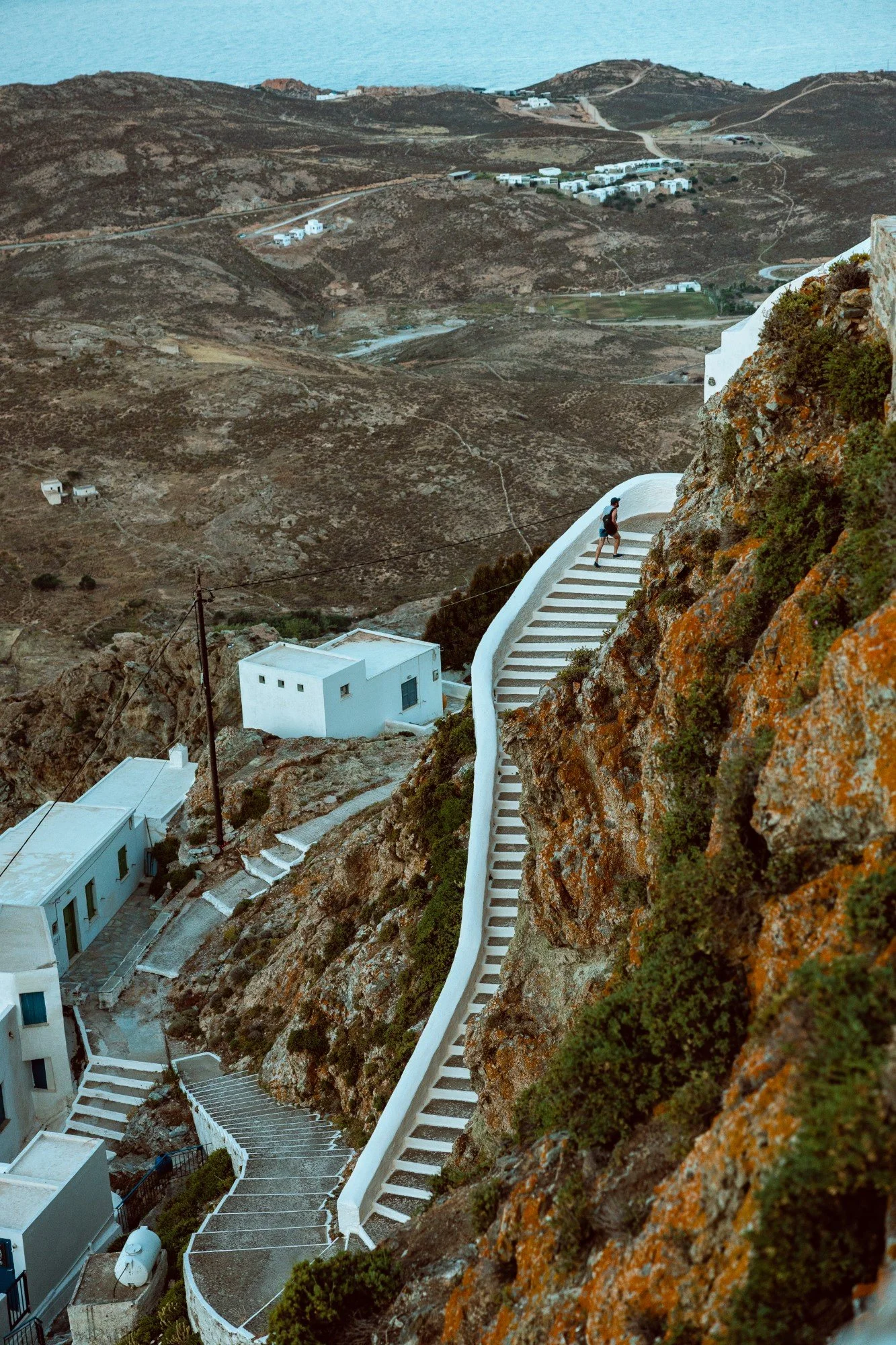 Steep hillside with a winding staircase, white buildings, and sparse vegetation, overlooking a body of water in the distance.