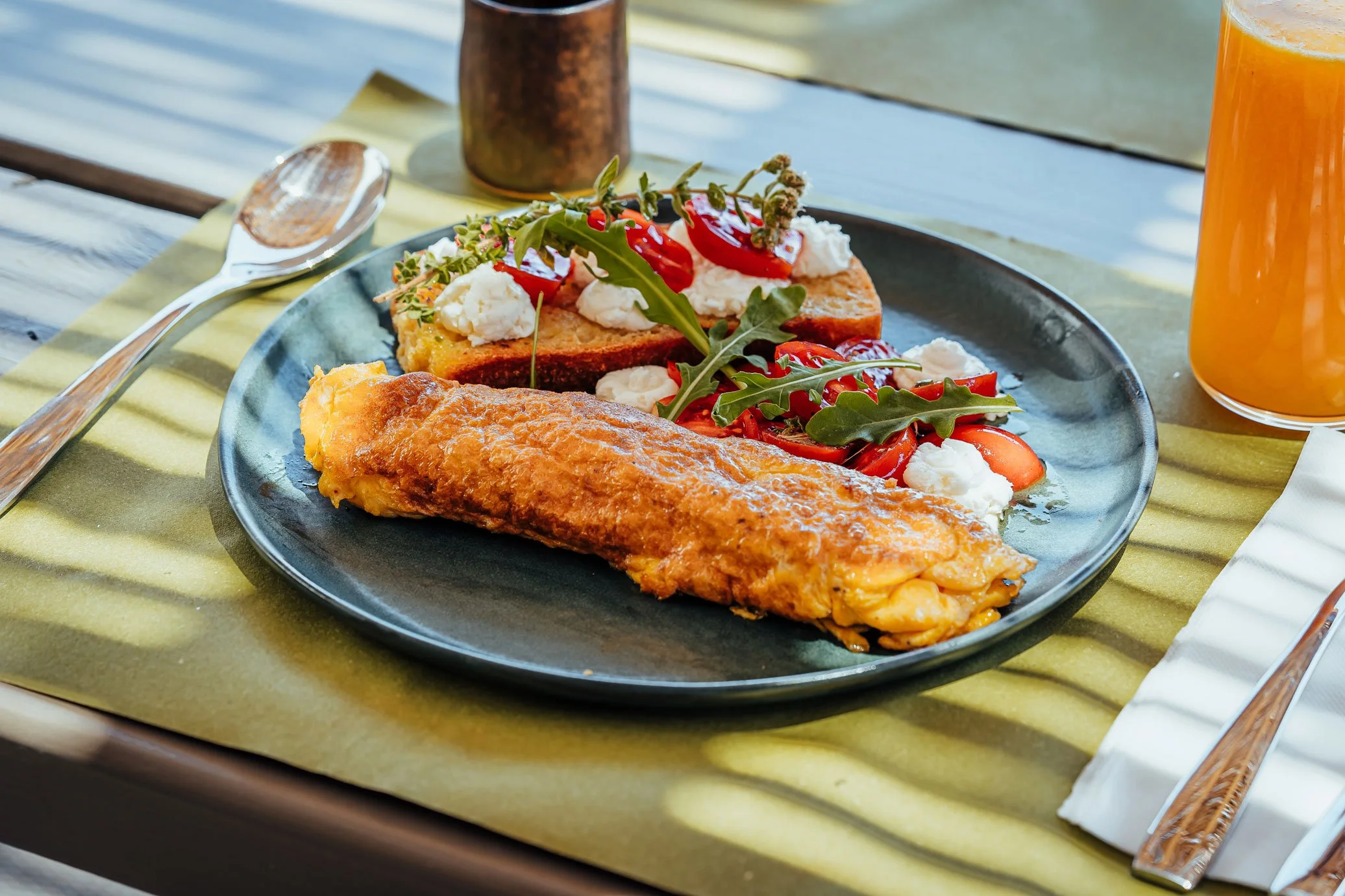Fried fish fillet served with a side salad of tomatoes, arugula, mozzarella, and bread topped with cheese and herbs, on a dark ceramic plate at a breakfast table.