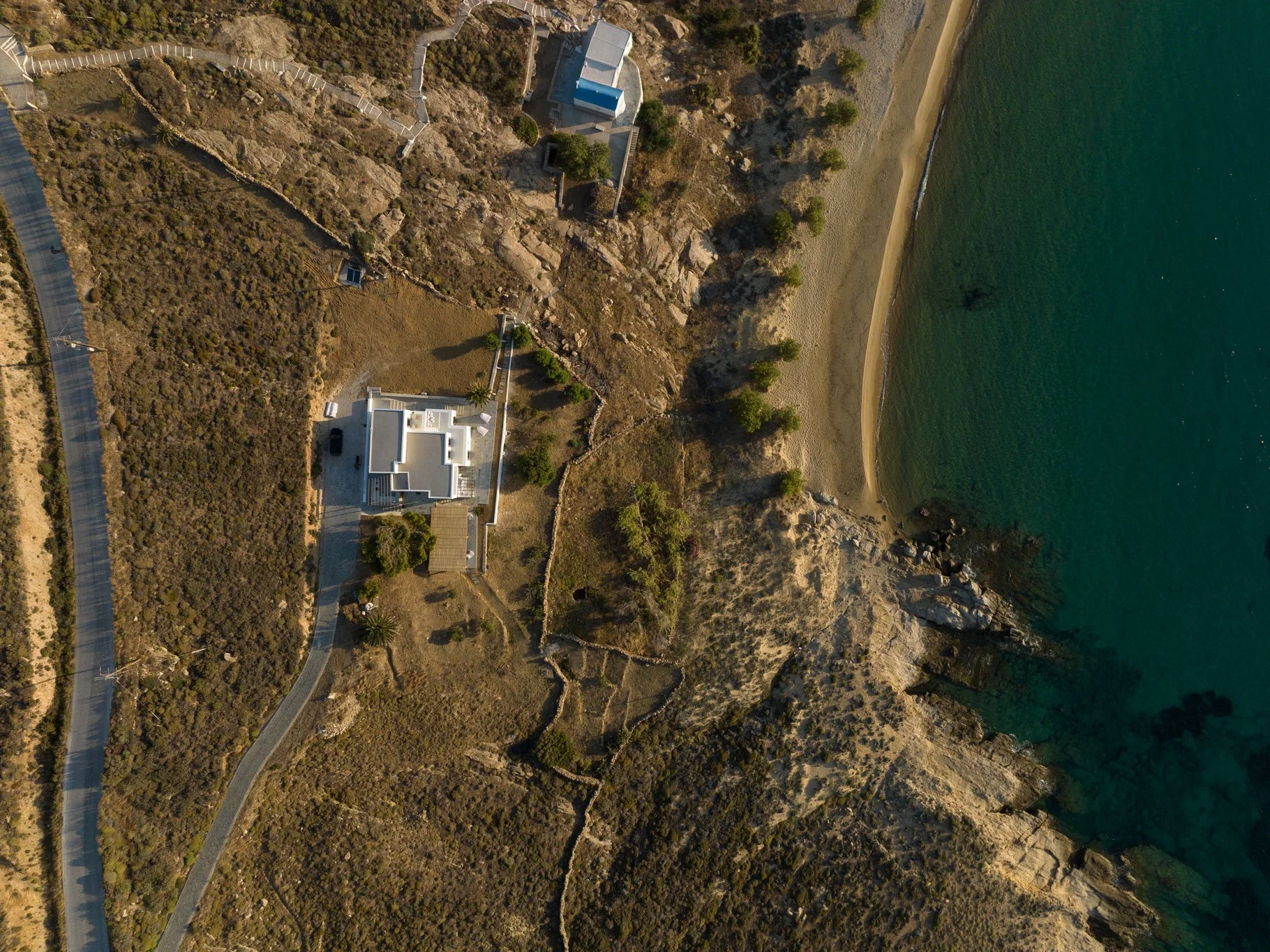 Aerial view of a coastal property with a white house, surrounded by a winding driveway, situated on a rocky hillside near a sandy beach with calm green water.