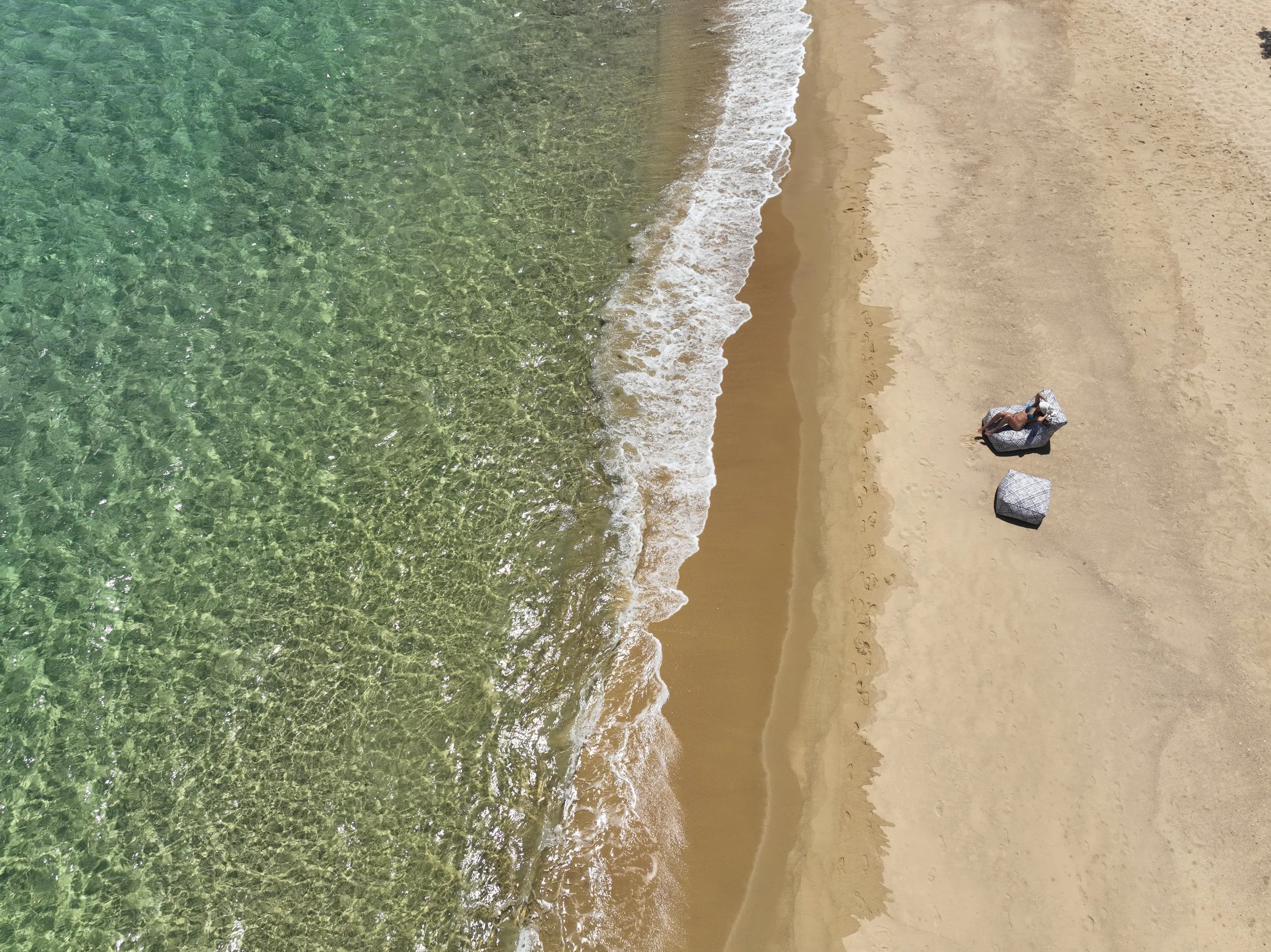 Aerial view of a sandy beach with clear green water and two lounge chairs and a person relaxing on the sand.