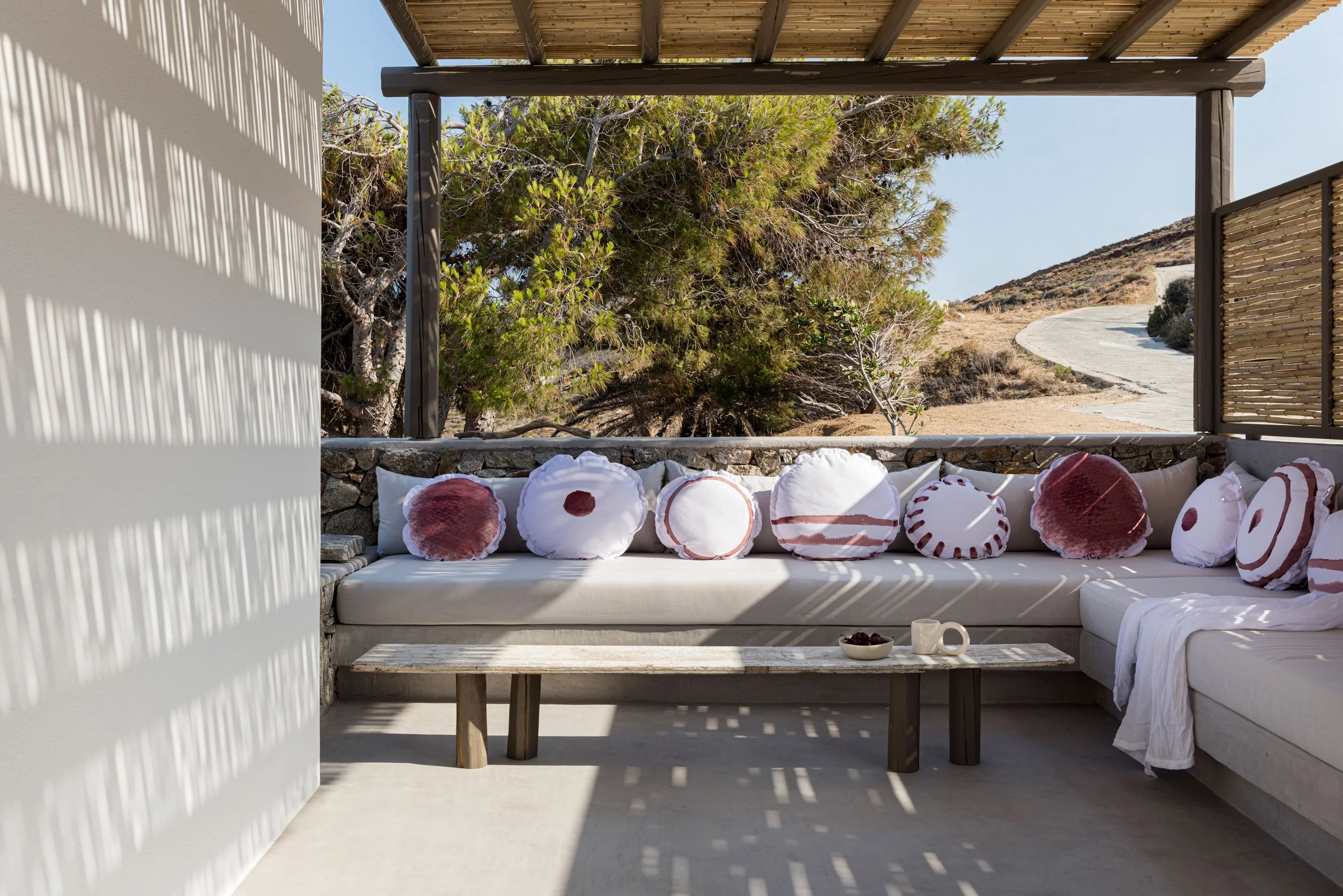 Outdoor seating area with white cushions and decorative pillows with red and white patterns, a coffee table with cups and a bowl of cherries, surrounded by a stone wall and overlooking a natural landscape with trees and a winding path.