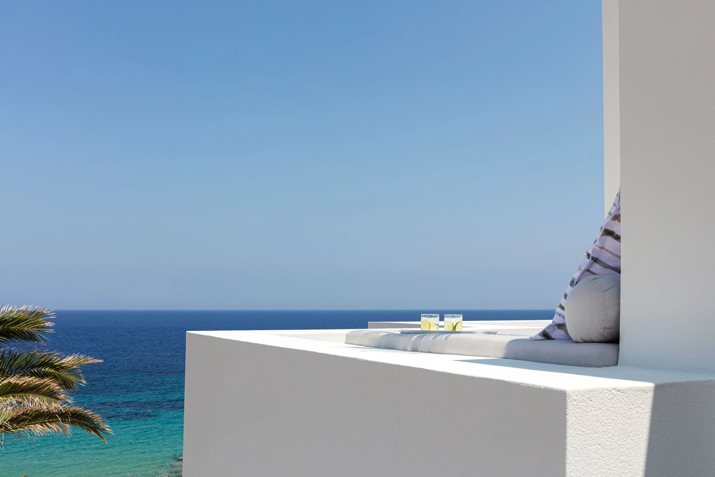 A white outdoor seating area on a balcony with two glasses of water and lemon slices overlooking the ocean under a clear blue sky.