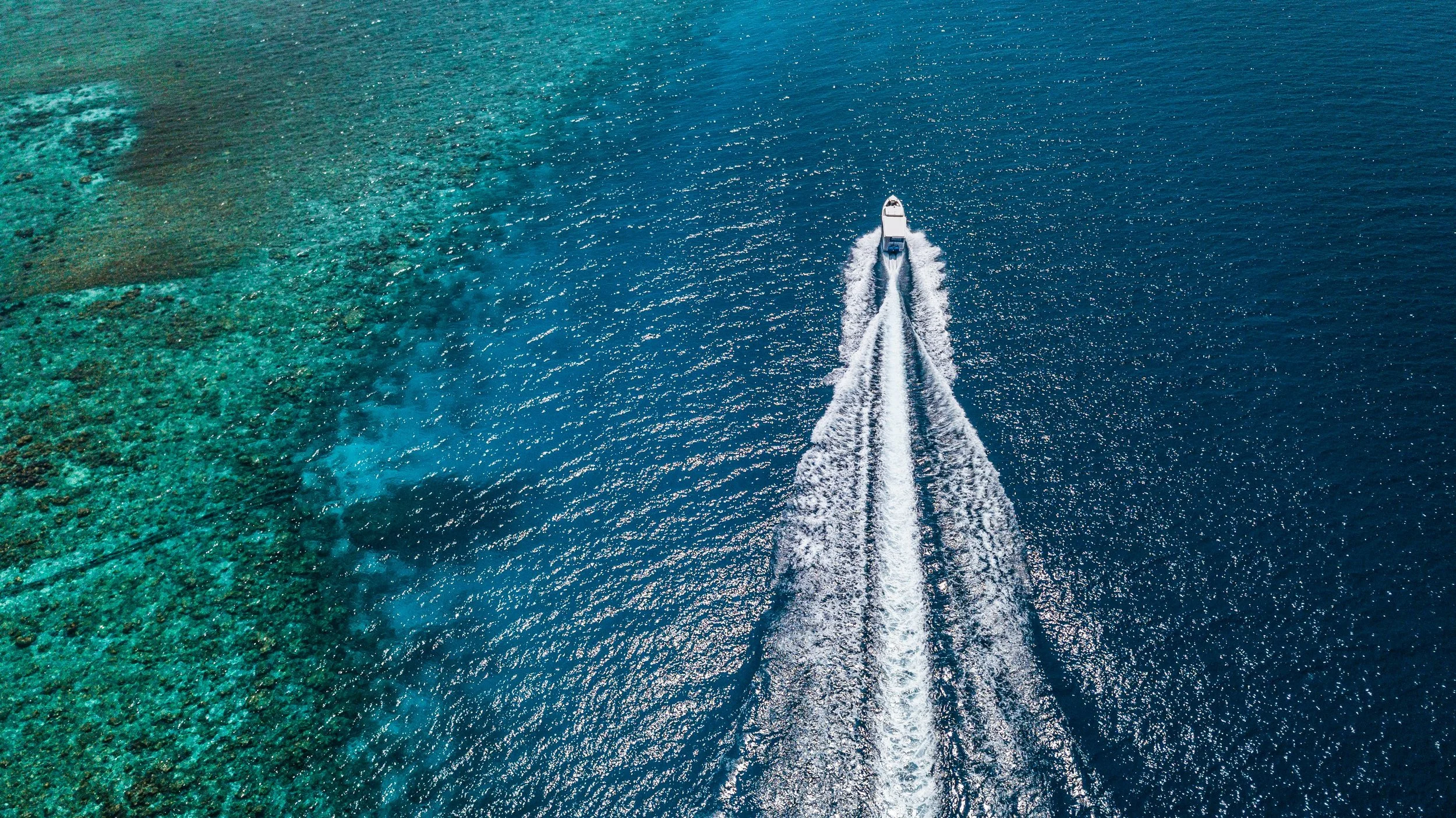 A white motorboat speeding across the ocean, creating a long wake, with vibrant turquoise coral reef on the left.