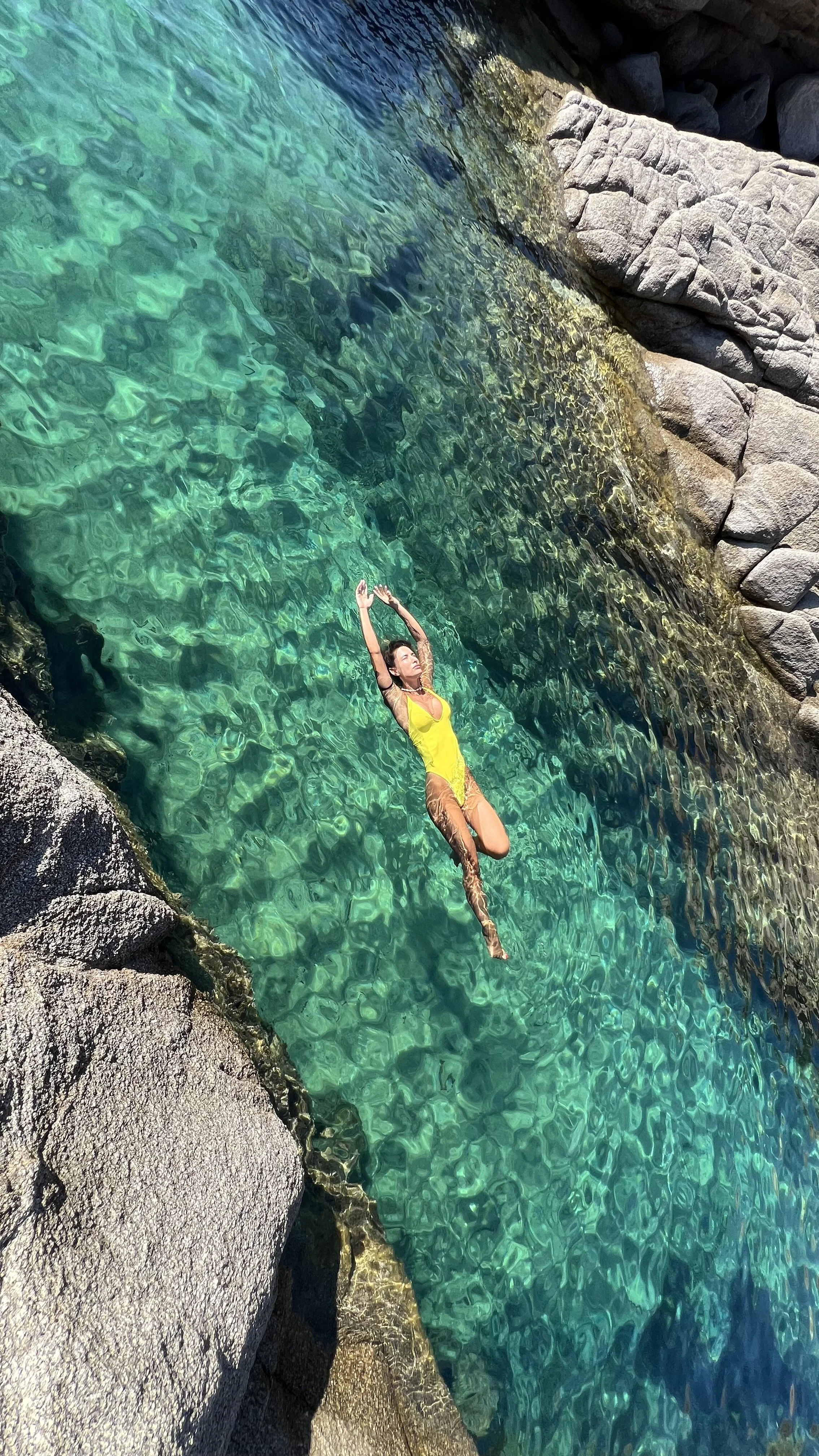 A woman in a yellow swimsuit floating on her back in clear turquoise water near rocks.