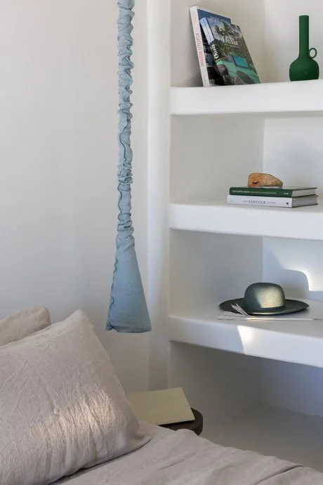 Part of a bedroom with beige bedding and pillow, white walls, a corner shelf with books, green vase, rock, gray hat, and a blue fabric hanging from the ceiling.