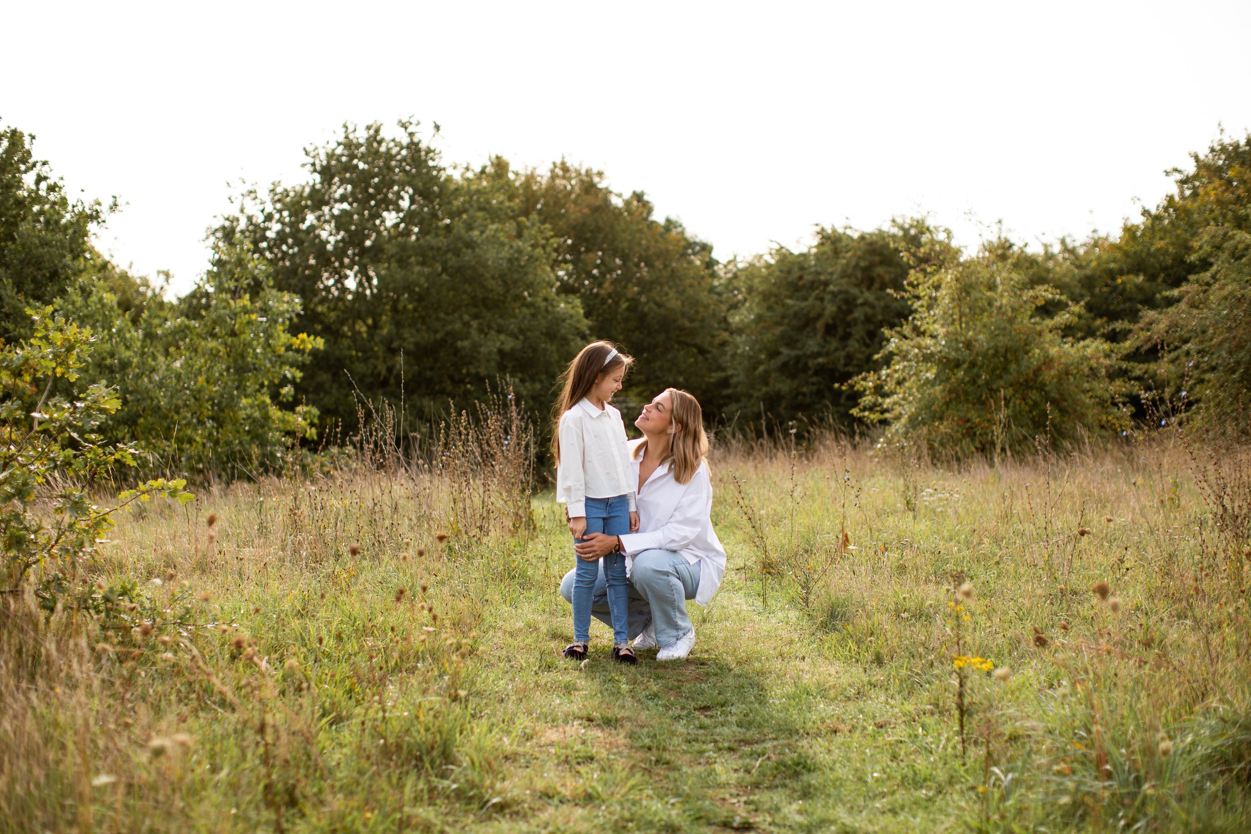 A woman kneeling in a grassy field, holding a young girl by the hands, with trees in the background.