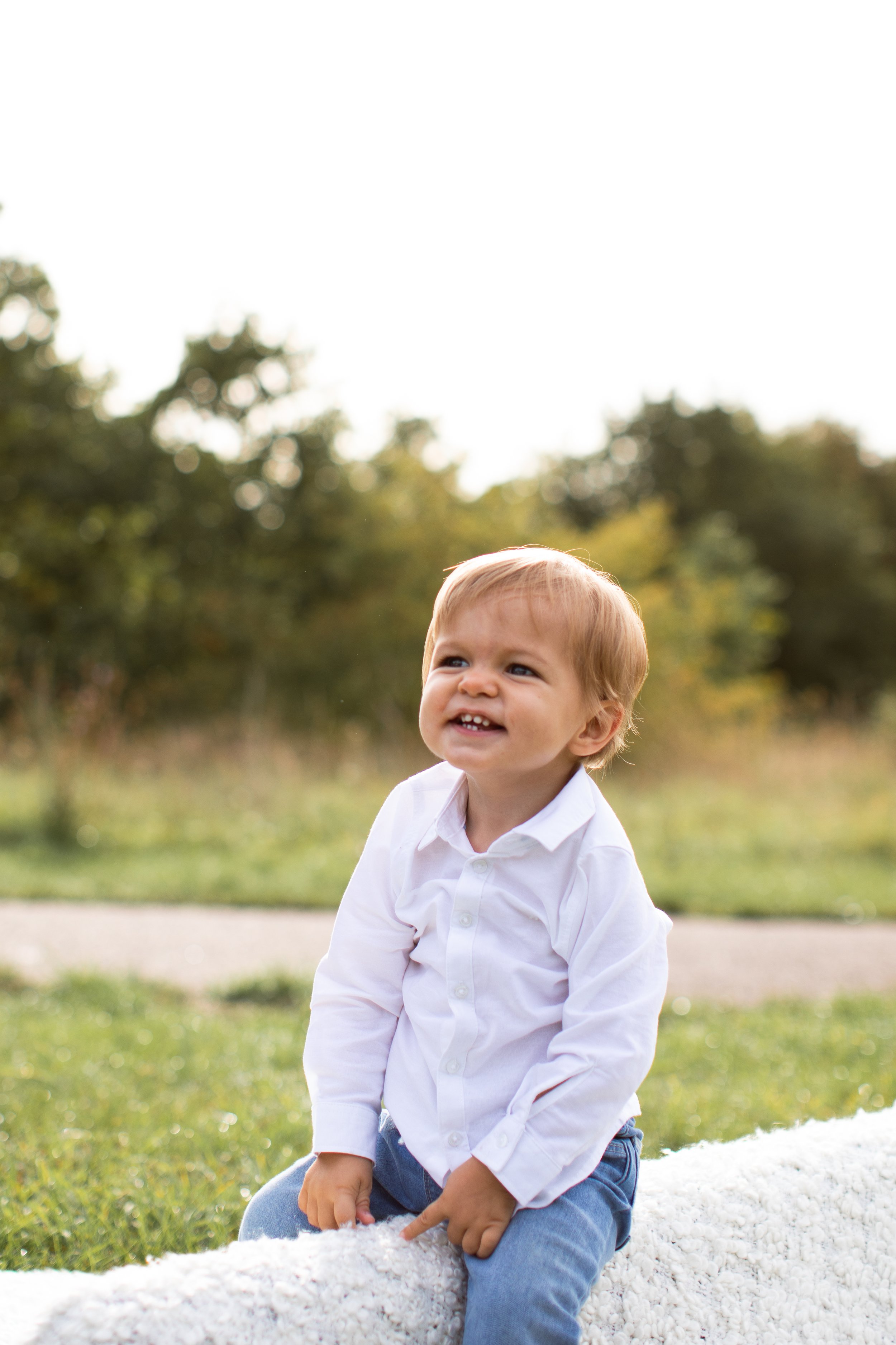 Young boy with light brown hair wearing a white shirt and blue jeans, sitting outdoors on a blanket, with trees in the background.