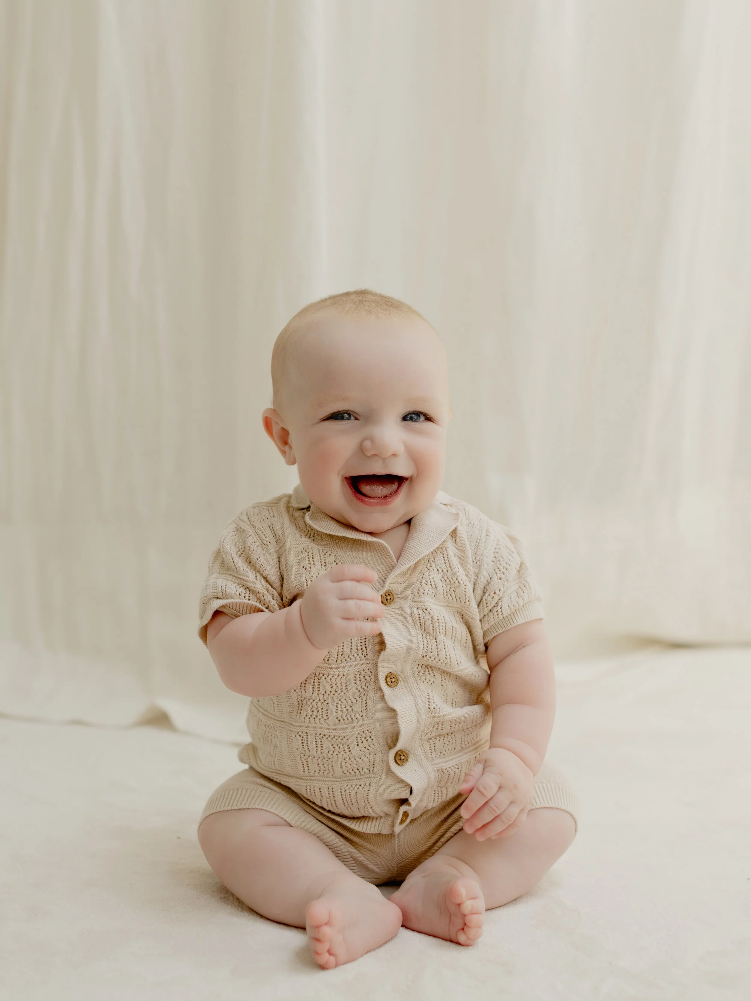 A smiling baby boy with brown hair sitting on a bed with white bedding and beige pillows.