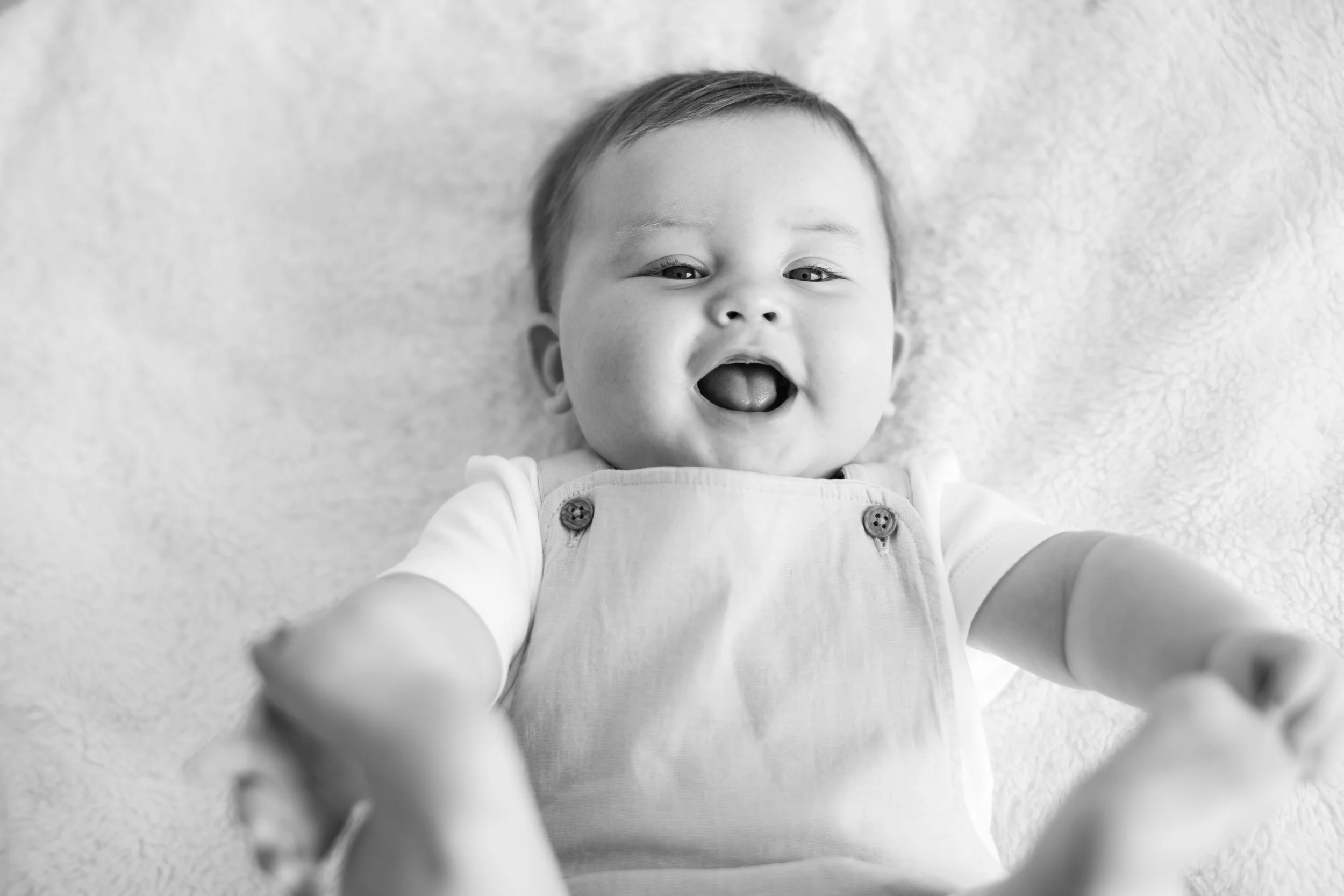 A smiling baby lying on a soft surface, wearing a light-colored shirt and overalls with buttons, reaching towards the camera.