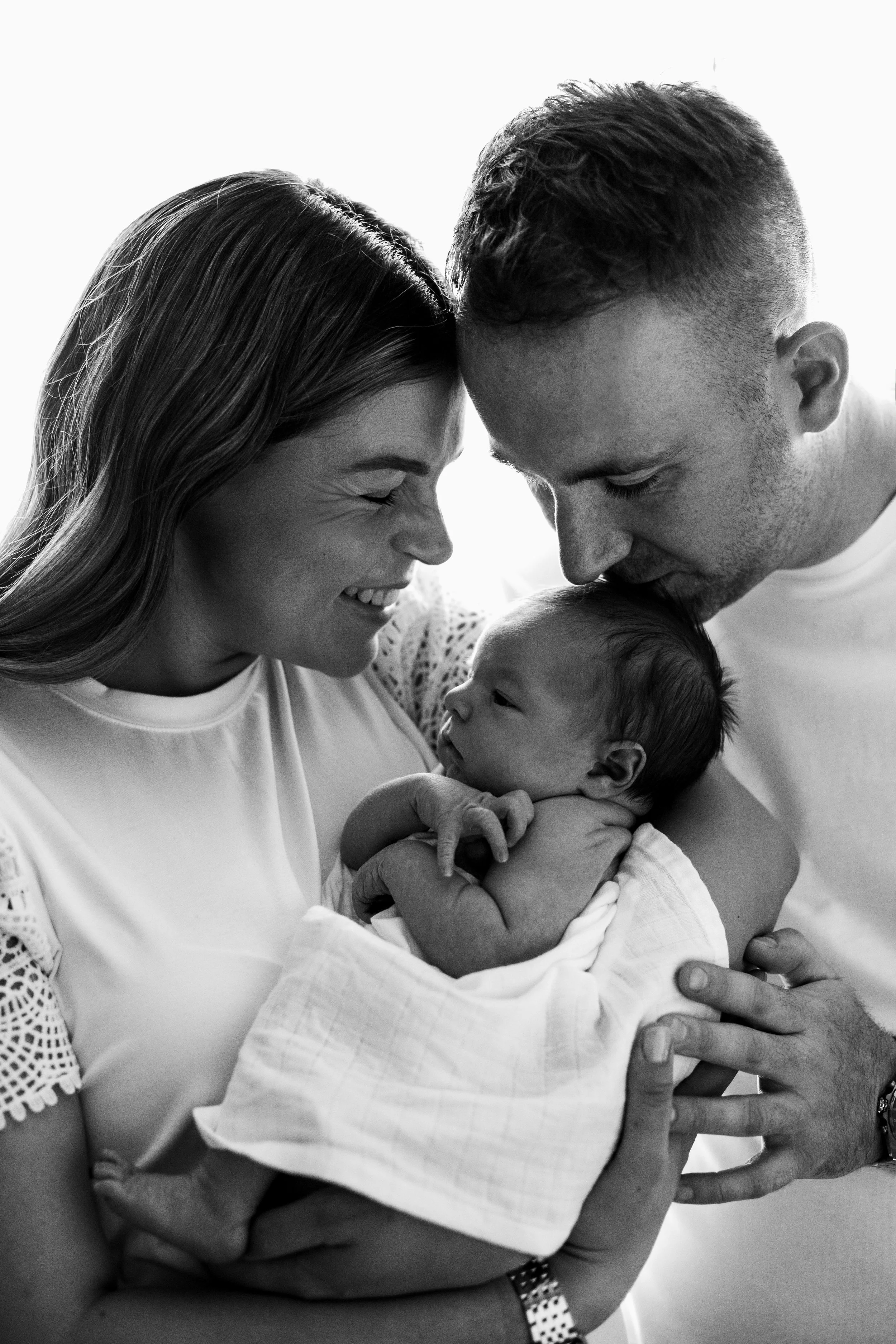 A black-and-white photo of a happy family of three, with a mother, father, and newborn baby. The mother and father are gently holding the baby, who is wrapped in a blanket, and they are touching their foreheads together, smiling and showing affection.