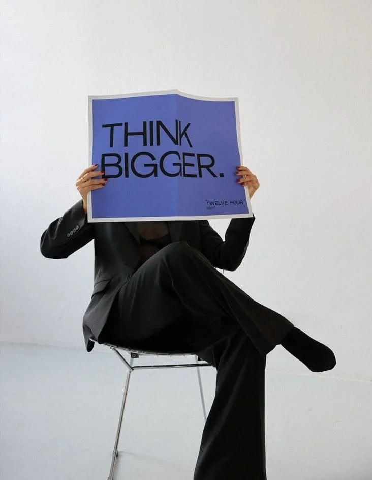 Women sitting on chair holding poster paper that says think bigger.