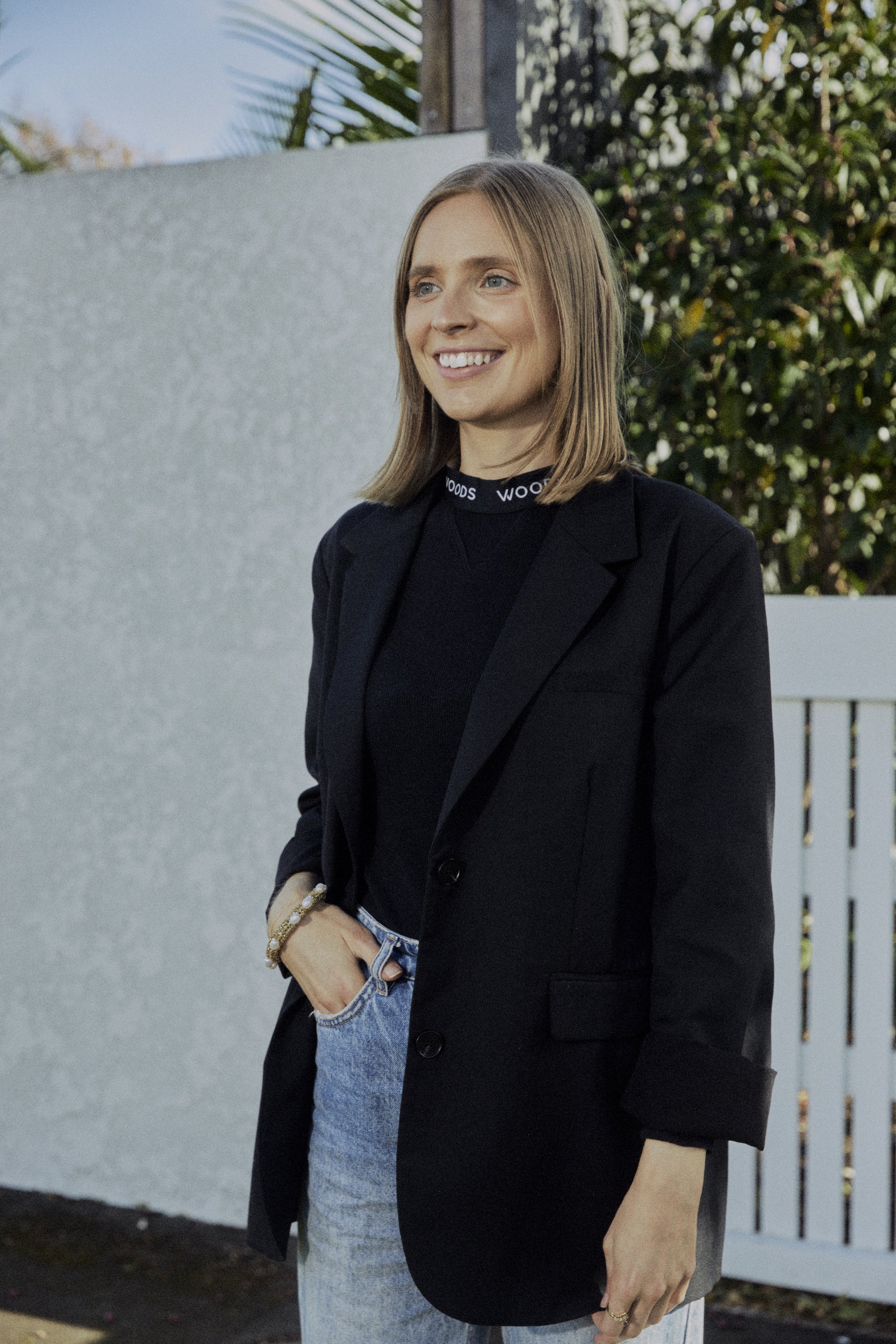A woman with shoulder-length light brown hair smiling outdoors, wearing a black blazer, black top with 'WOODS' written on the collar, and light blue jeans.
