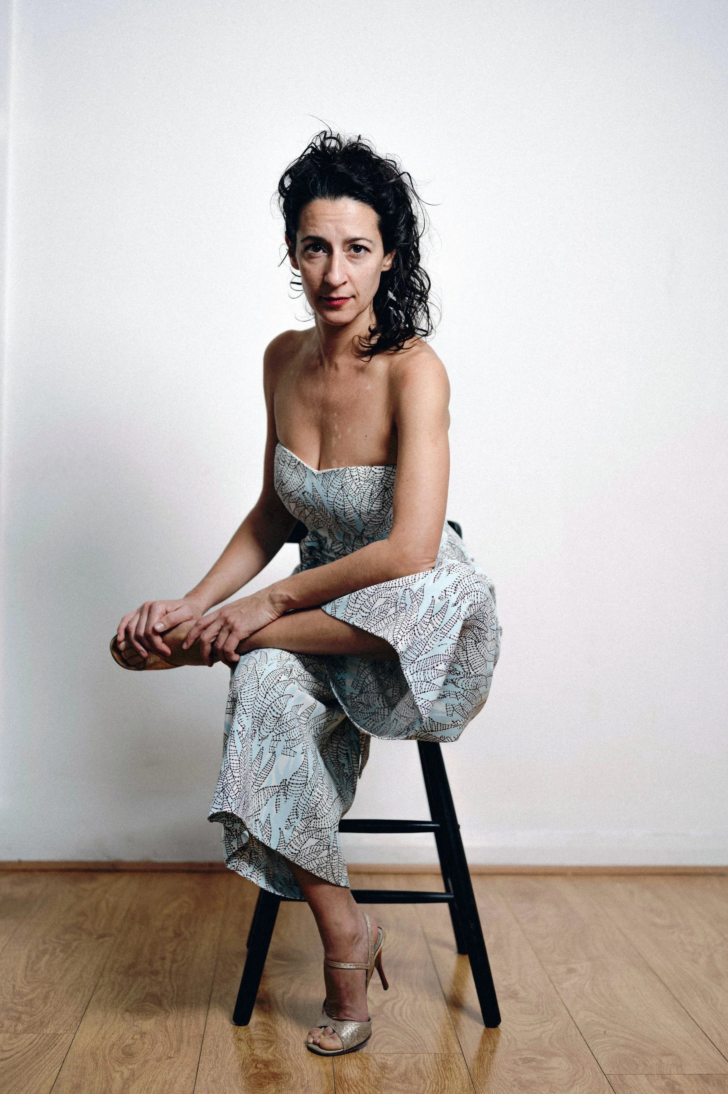A woman with dark, curly hair seated on a black stool against a white wall. She is wearing a strapless, patterned jumpsuit and high-heeled sandals.