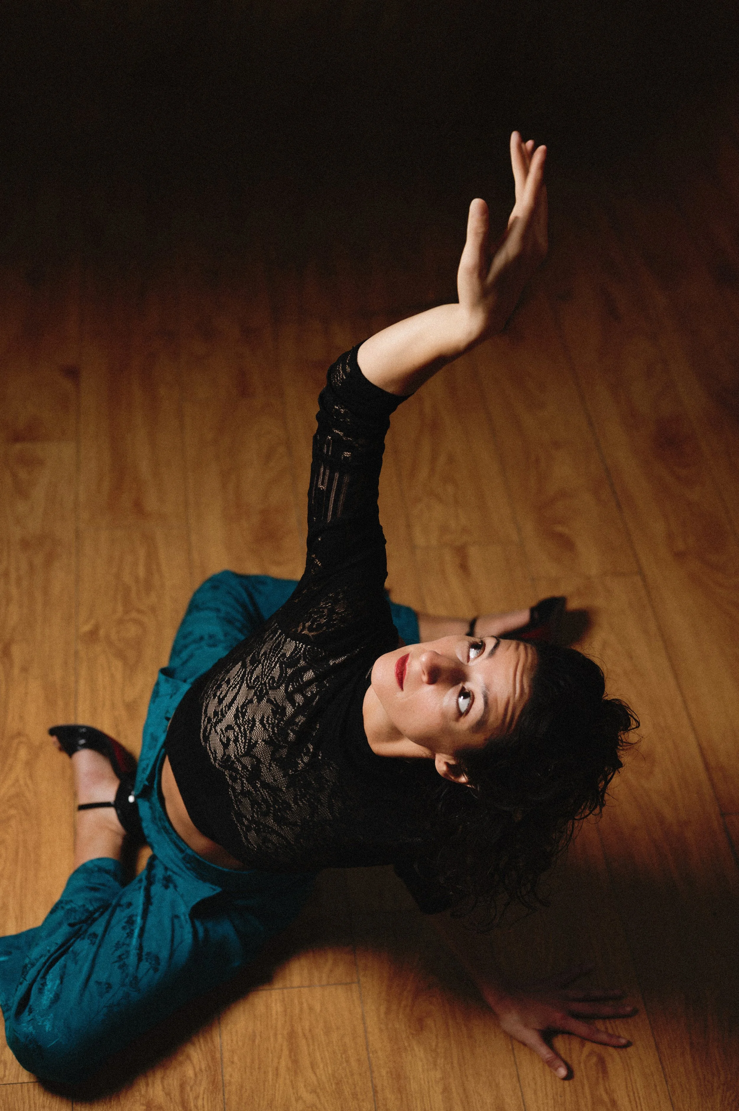 Woman with curly hair and red lipstick lying on a wooden floor, reaching upward with her right arm extended and wearing a black lace top and teal pants.
