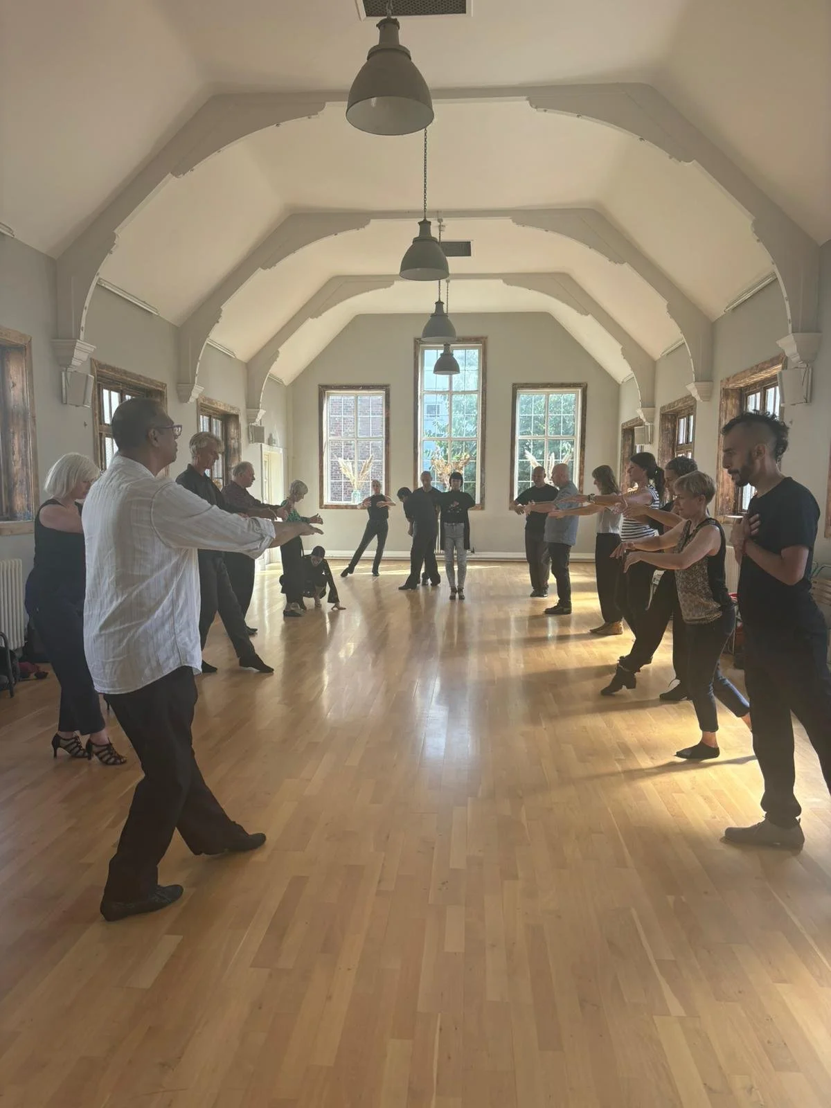 Group of people practicing partner dance in a spacious room with high arched ceiling and large windows.