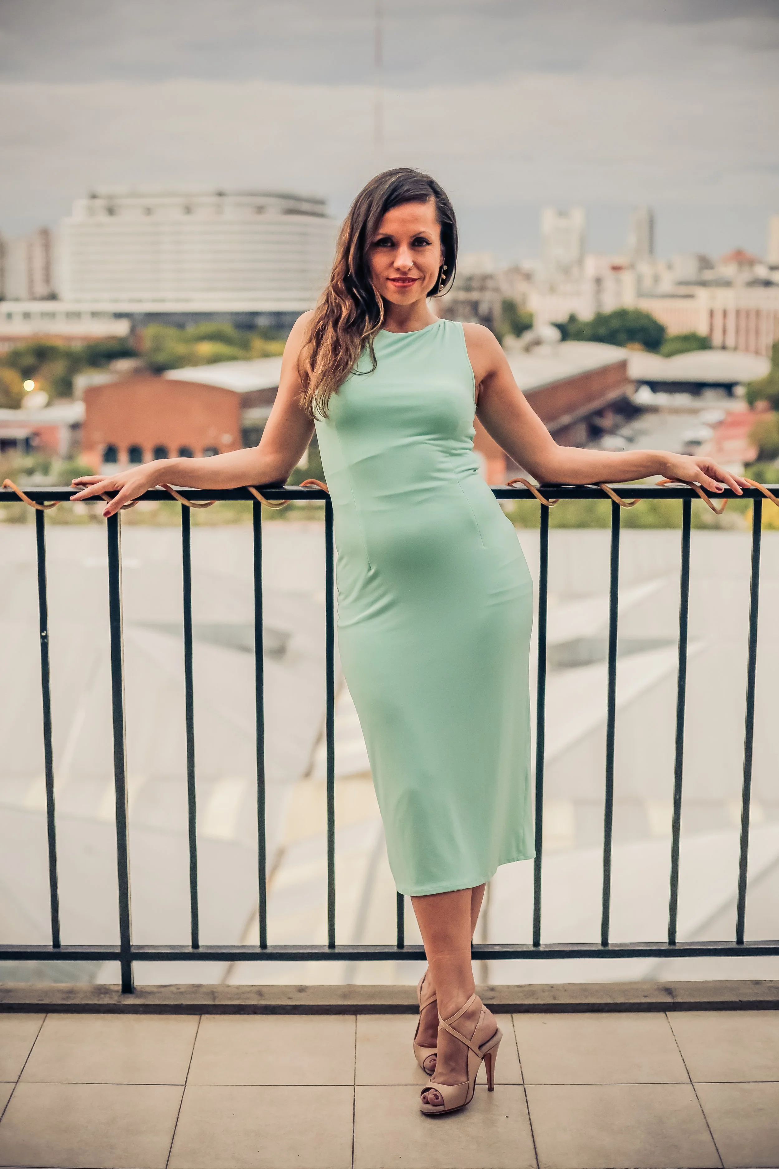 A woman in a light green dress and beige high heels standing on a balcony with a cityscape in the background.