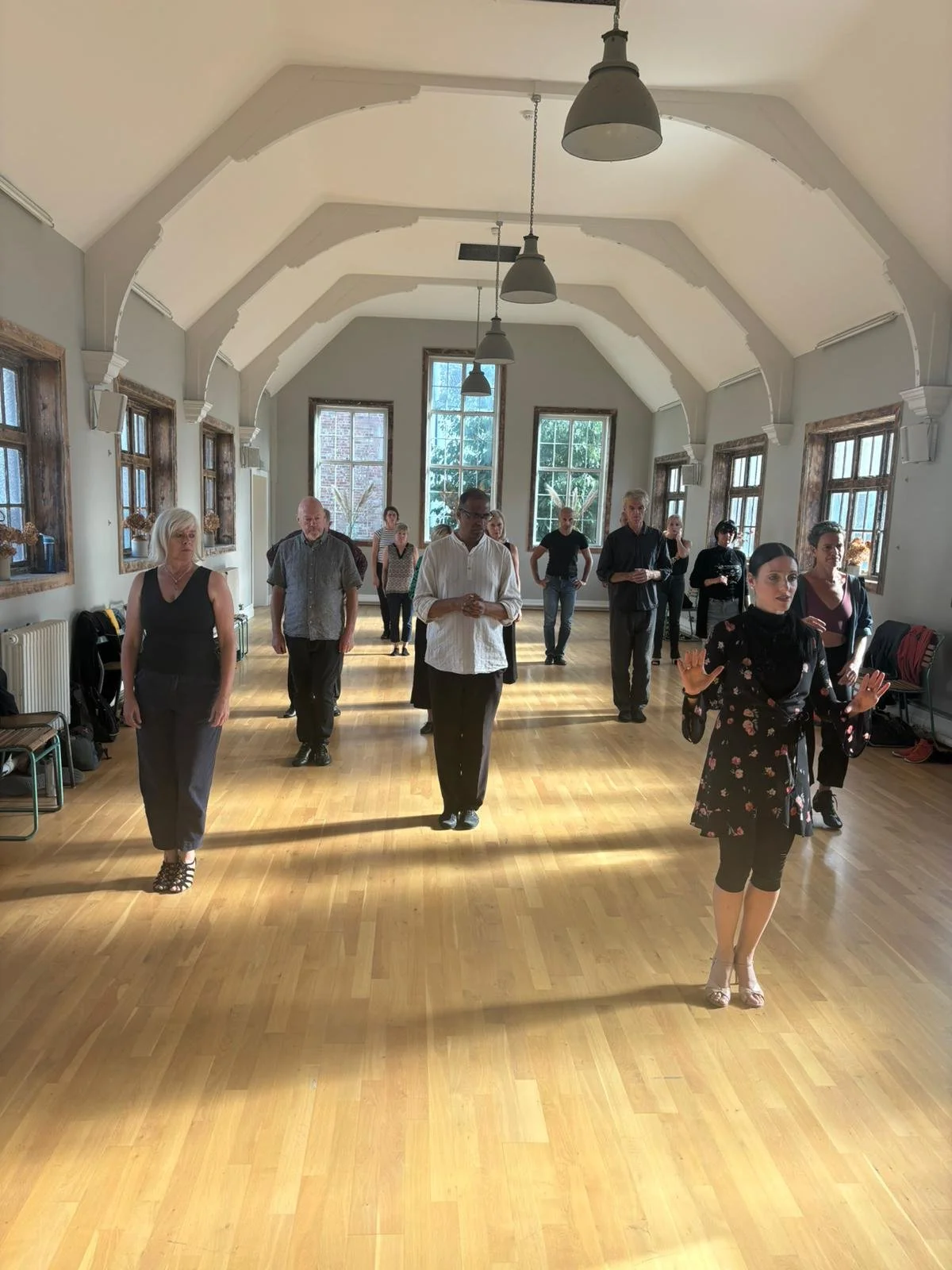 People participating in a dance or movement class in a spacious room with large windows, wooden floors, and high ceilings with hanging lights.