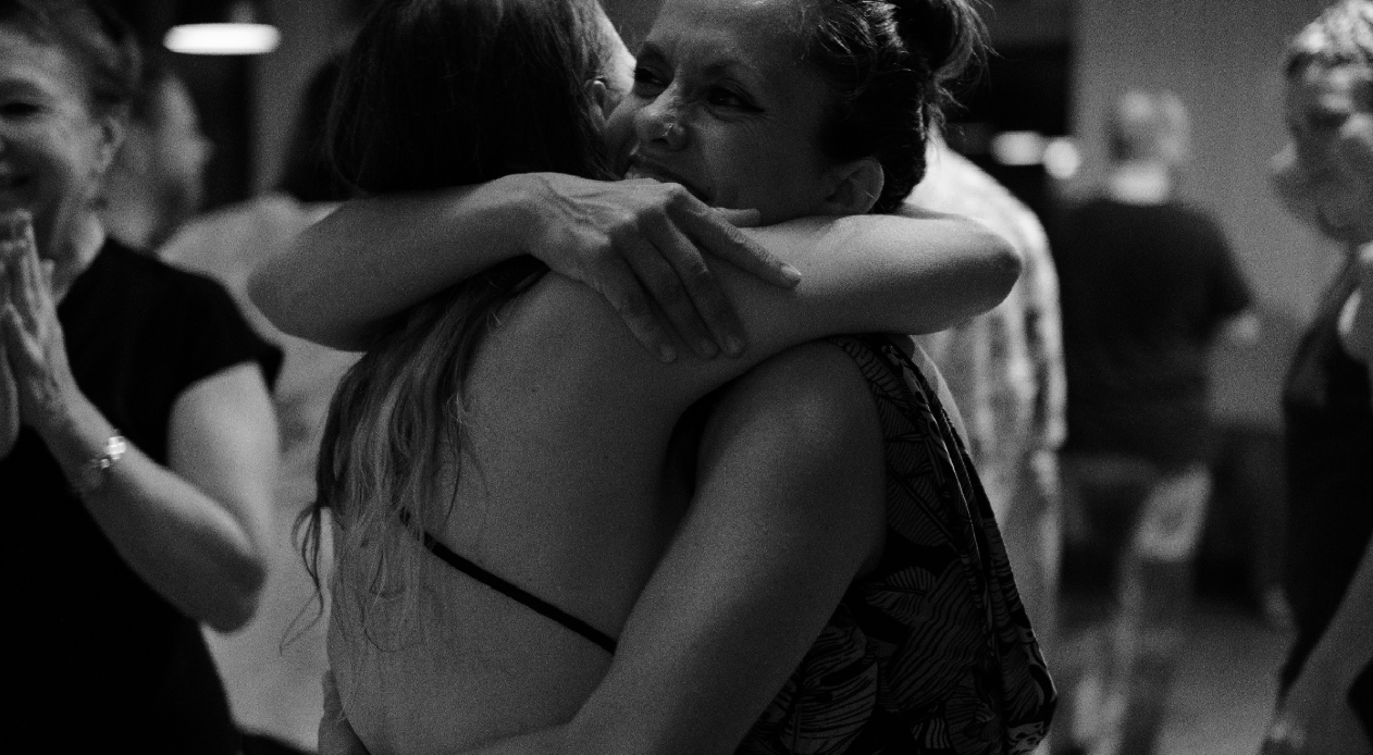 Two women embracing and smiling at each other in a social setting, with other people in the background.