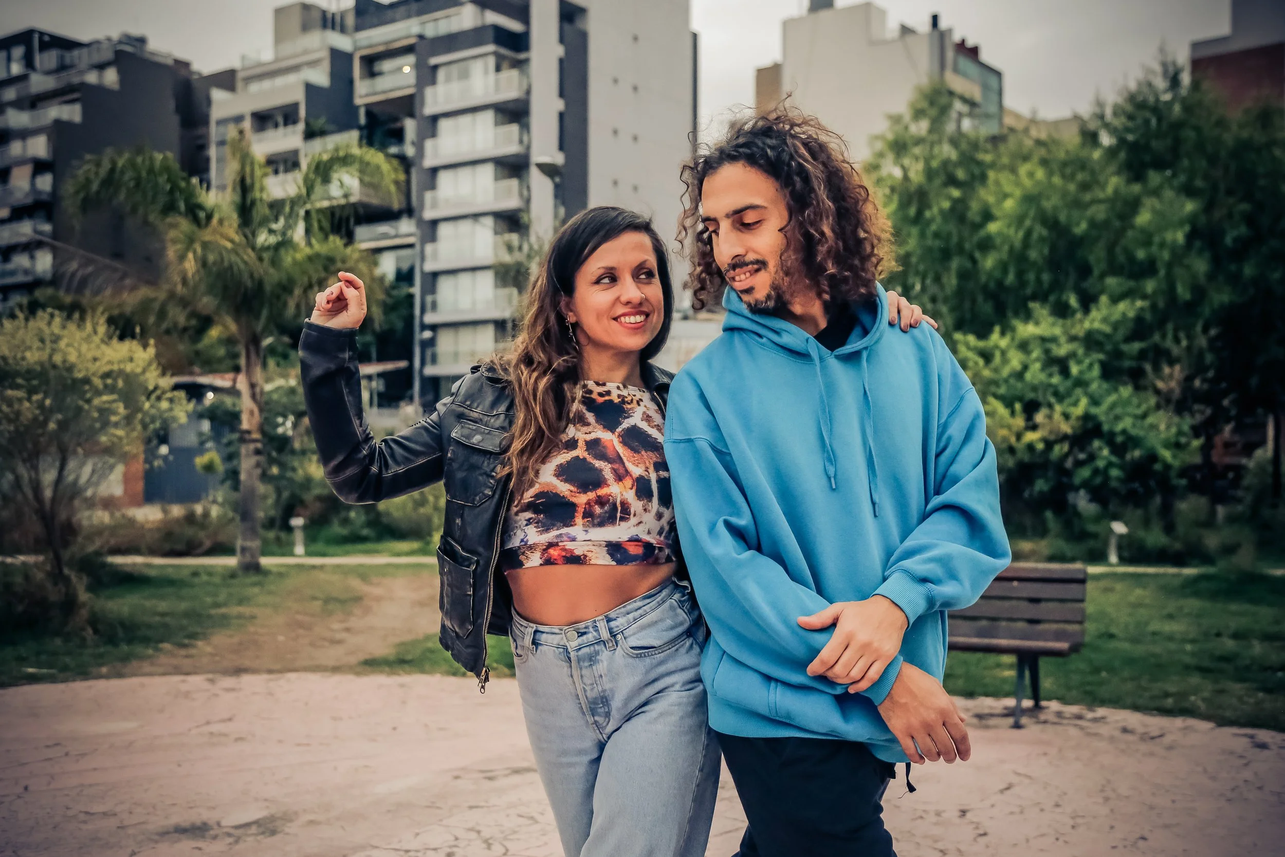 A woman and a man walking outdoors in a park in a city. The woman is smiling and showing her bicep, wearing a leather jacket, animal print crop top, and jeans. The man is looking down, wearing a blue hoodie, with his arms crossed.