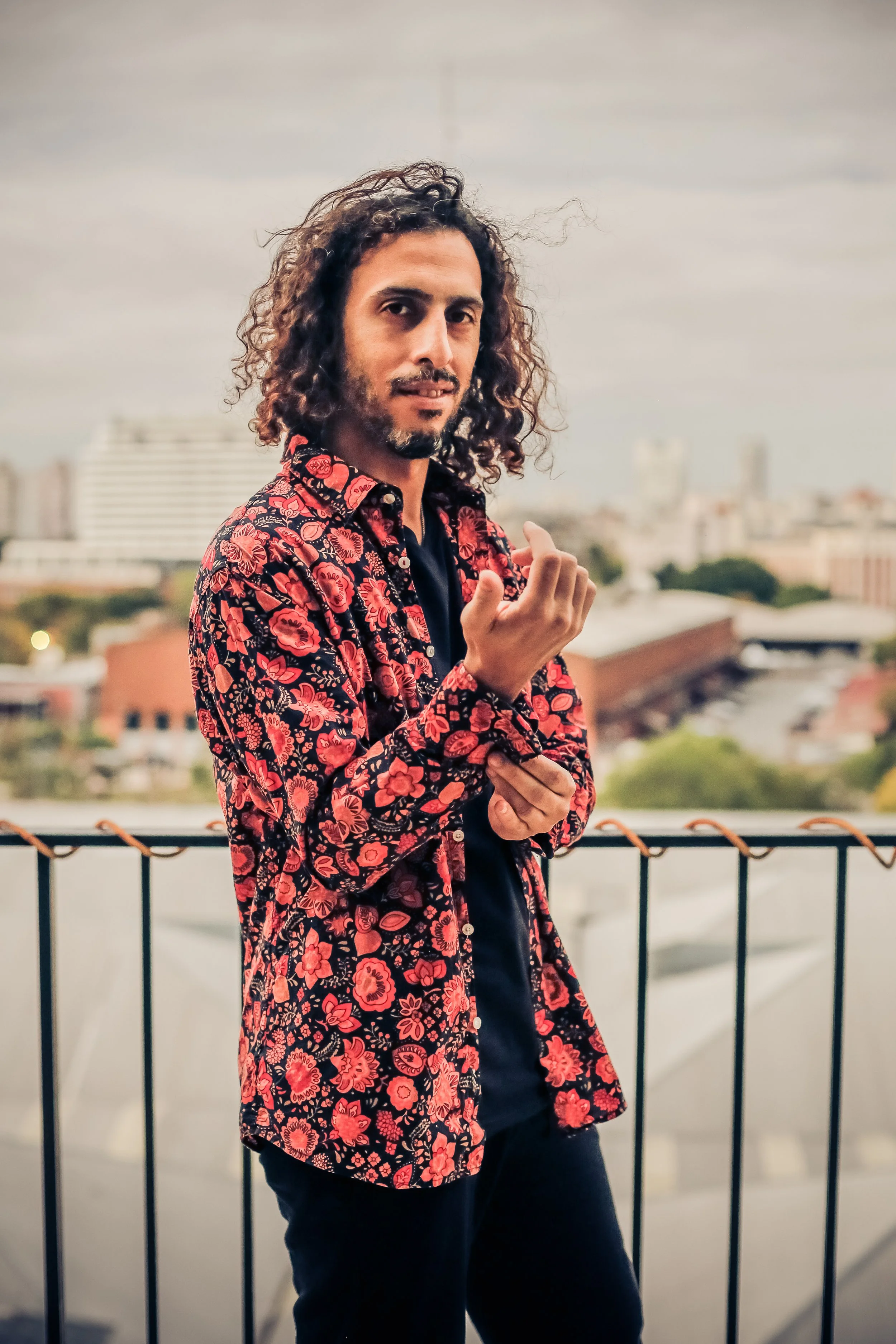 A man with curly hair and a goatee in a floral red and black shirt poses outdoors on a balcony with a cityscape in the background.