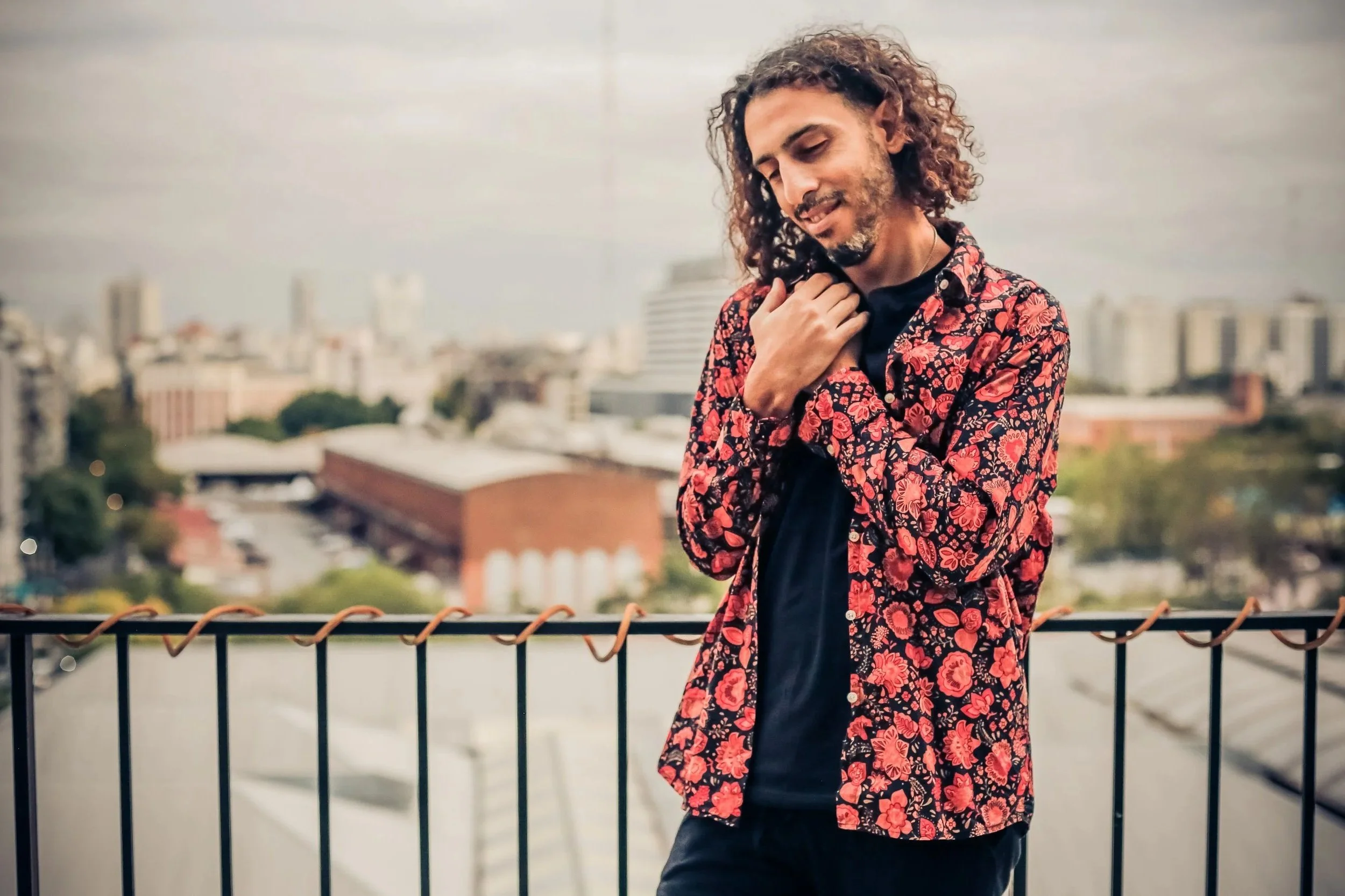 A man with curly hair and a beard wearing a red floral shirt over a black T-shirt standing on a rooftop with a cityscape in the background.