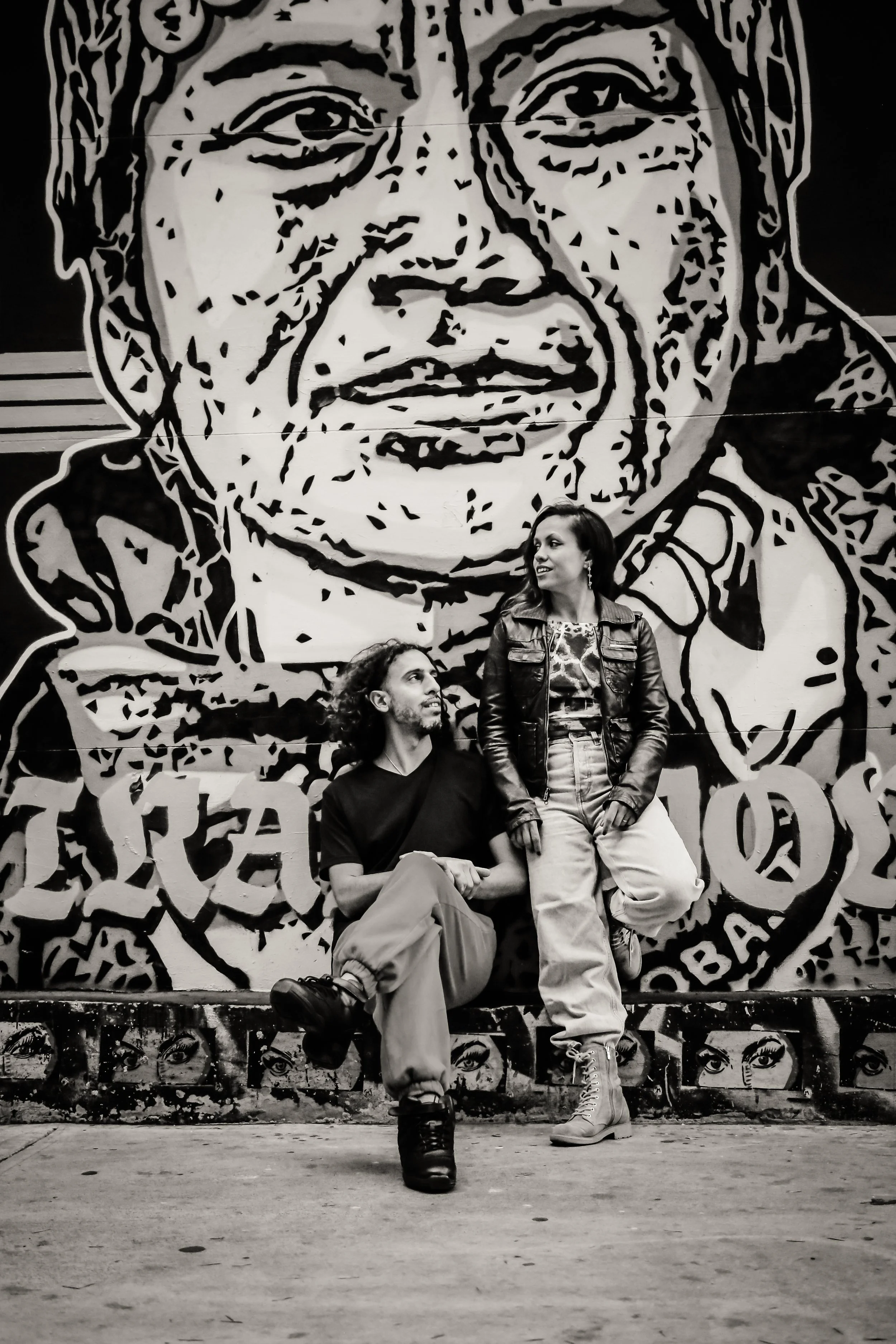 Two people, a man and a woman, sitting and standing in front of a large black-and-white mural of a woman's face with artistic eye designs at the bottom.