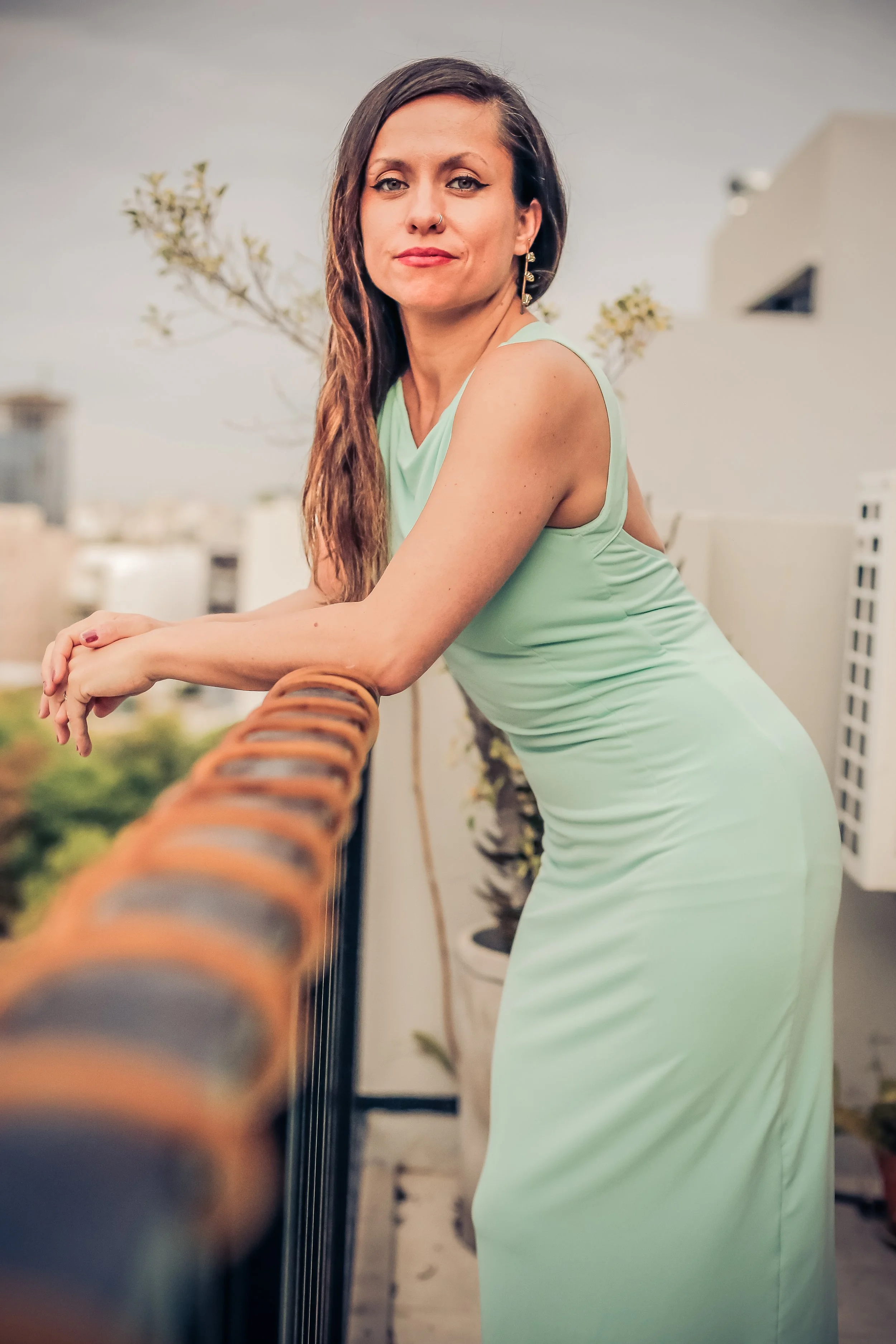 A woman with long brown hair, wearing a light green sleeveless dress, leaning on a balcony railing with city buildings and trees in the background.