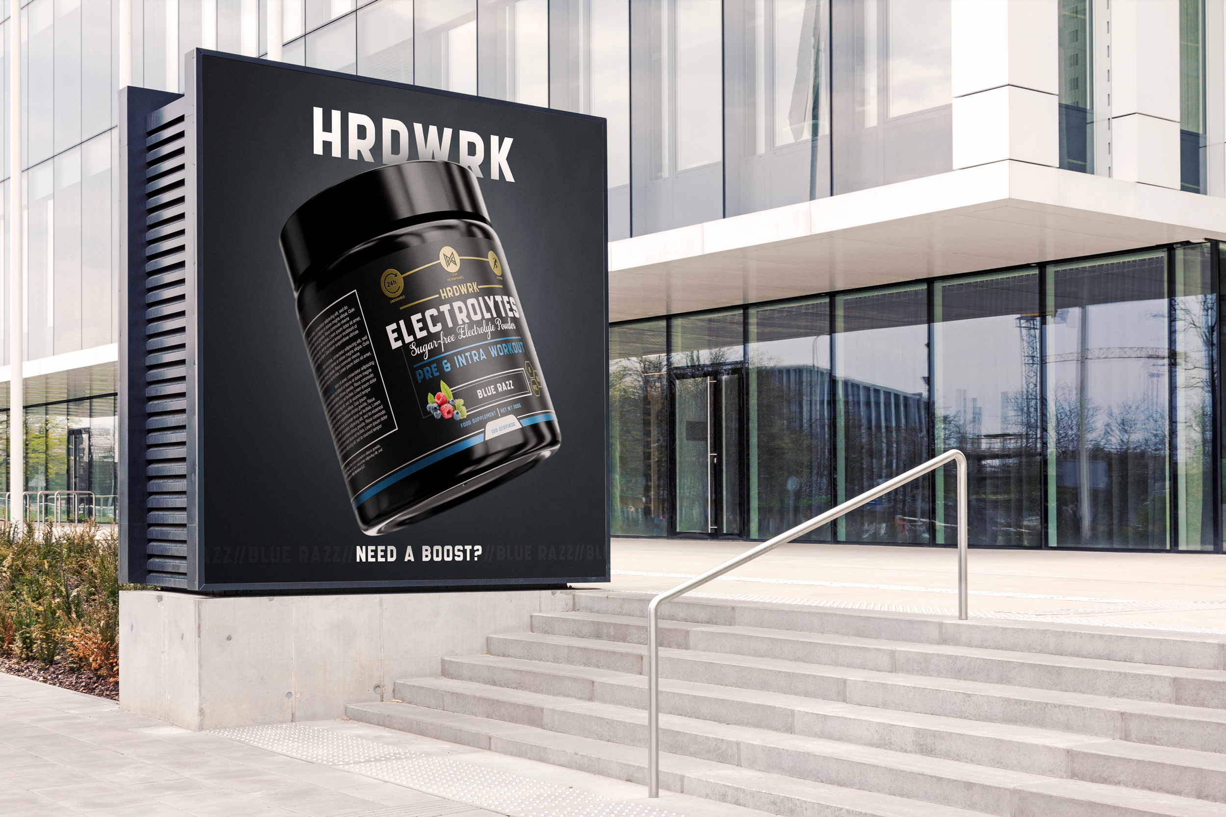 Digital billboard outside modern office building displaying a large container of HARDWRK Electolytes powder, with the text "Pre & Intra Workout" and "Blue Razz" flavor, along with the tagline "Need a Boost?"