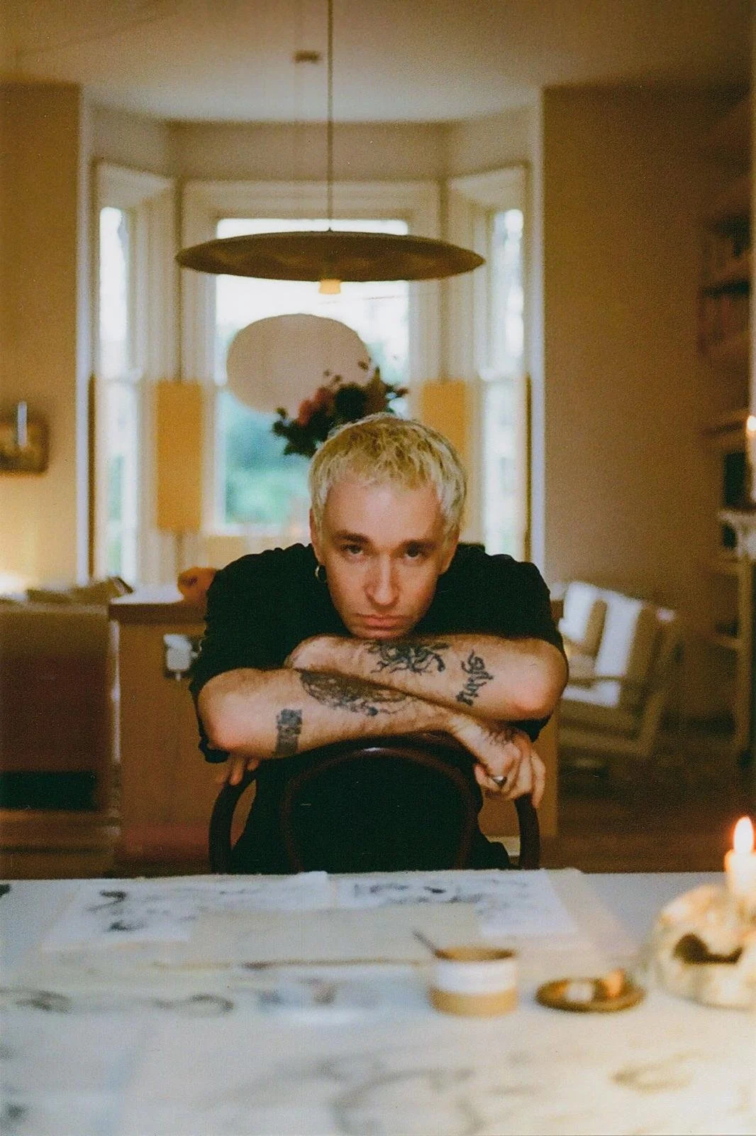 Ted Rogers, a non-binary person with short blonde hair and tattoos on their arms, leaning on a chair with arms crossed, sitting at a table in a cozy, warmly lit room with a large bay window and a hanging light fixture.