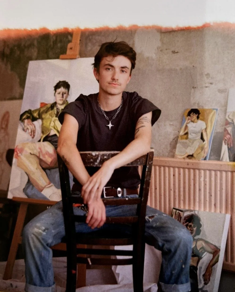 Brogan Bertie, a young man with dark hair and tattoos sitting on a chair in an artist's studio, surrounded by paintings of people, with a concrete wall background.