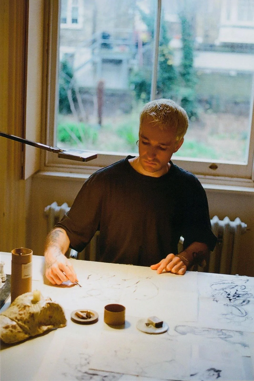 A young non binary person with blond hair and tattoos sits at a table by a window, drawing or writing on a large sheet of paper with various small objects and containers on the table.