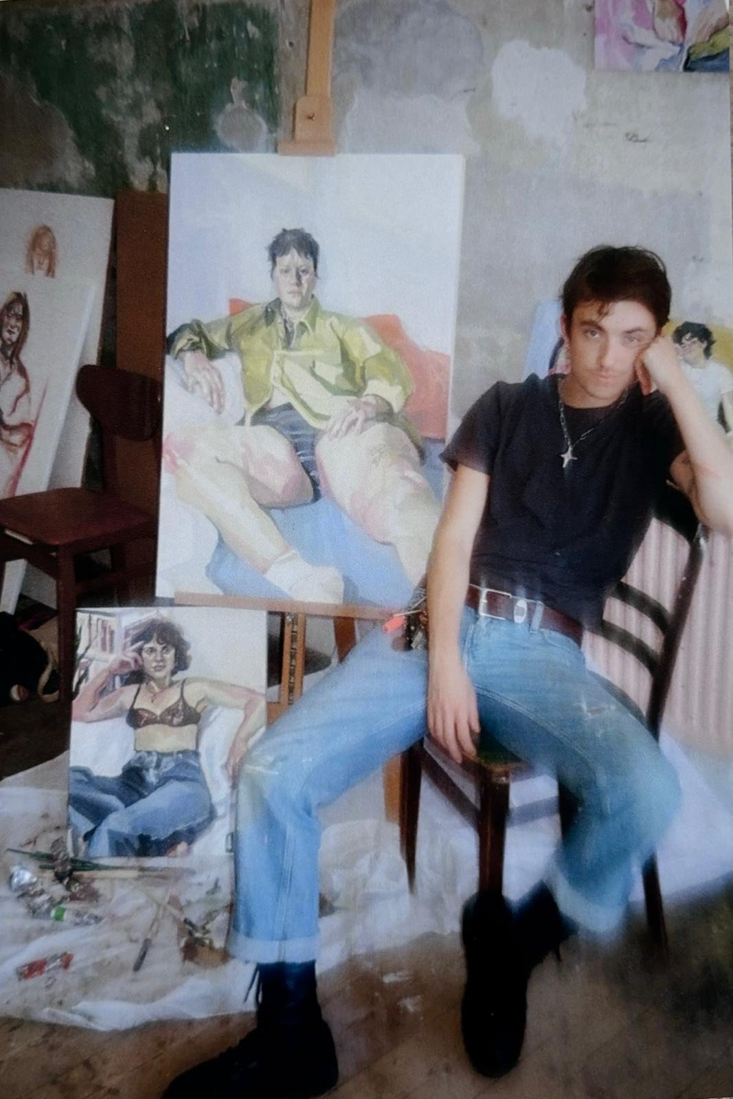Young woman sitting on a chair next to several completed paintings of fashion models, in an art studio with unfinished walls.
