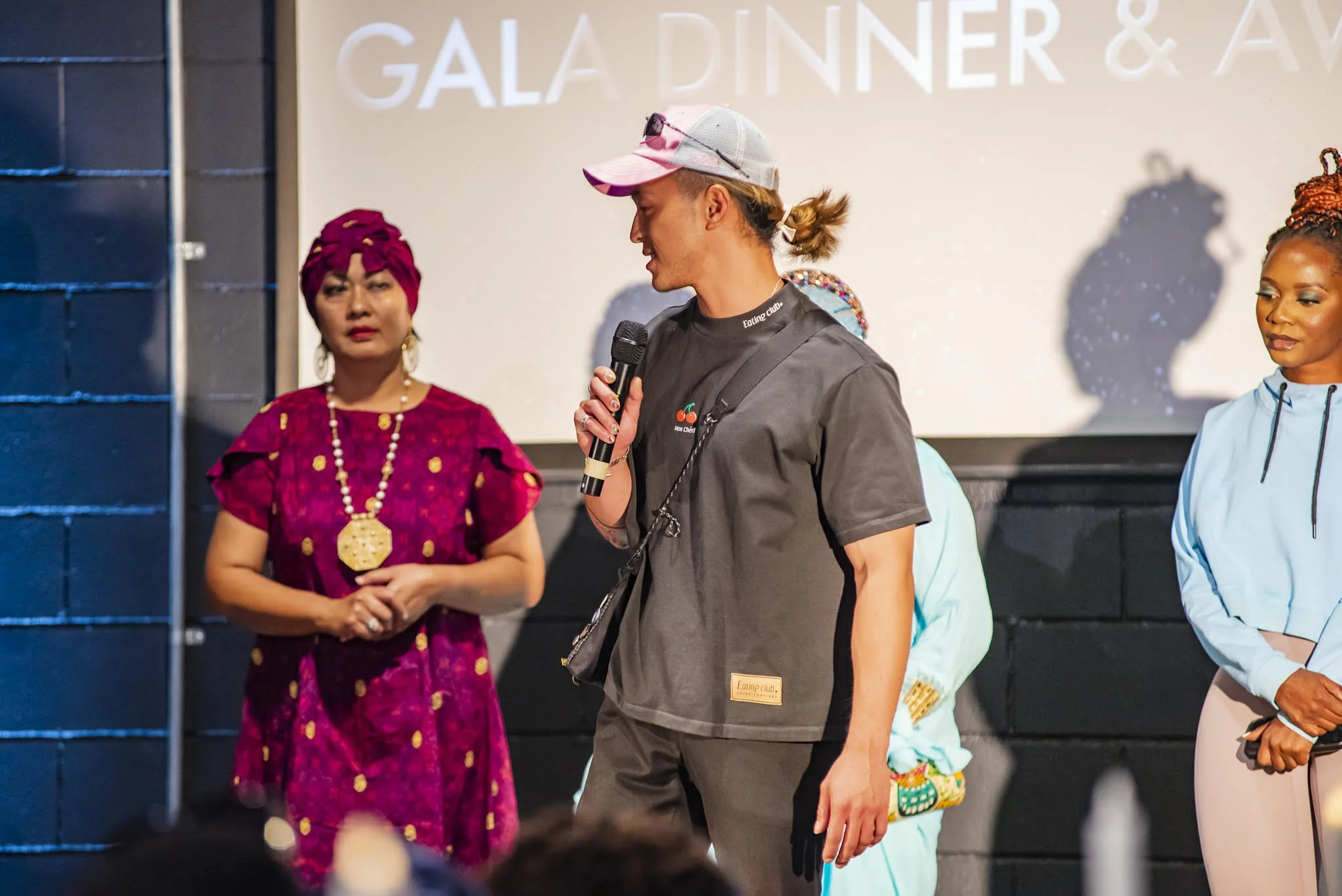 A group of diverse people standing on stage at an event, with a man holding a microphone and speaking. The background displays large text that reads 'GALA DINNER & A...' and people are dressed in colorful clothing.