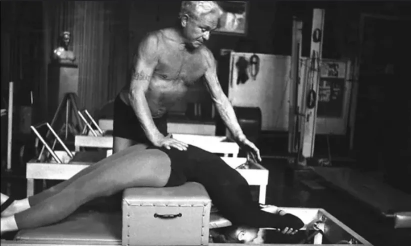 Joseph Pilates providing pilates exercices to a woman lying face down on a treatment table, in a room with Pilates equipment and framed pictures.