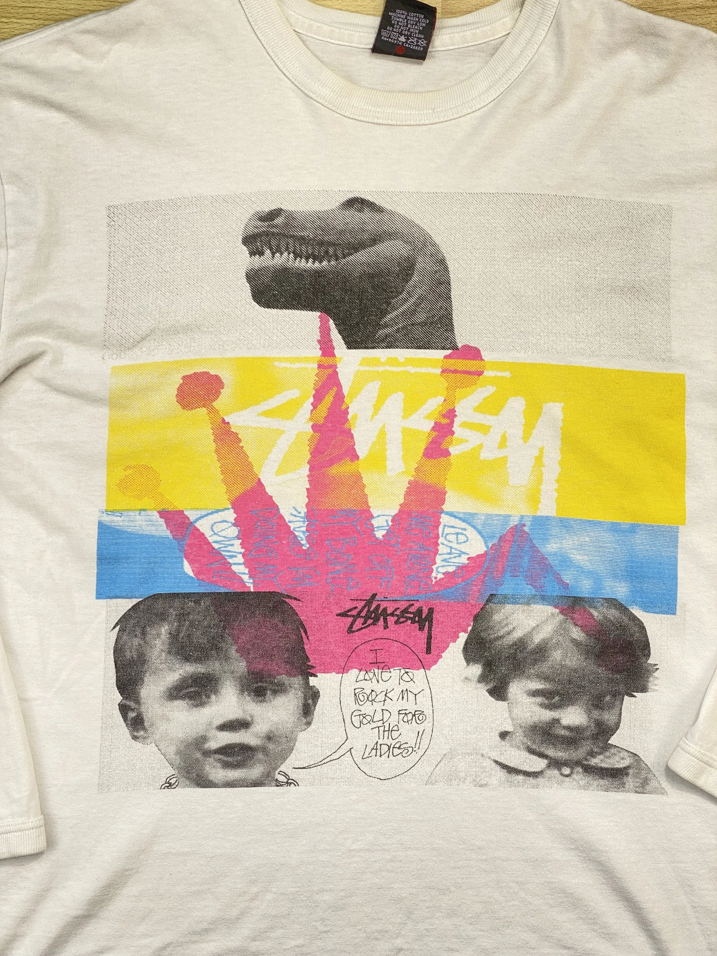 A graphic T-shirt featuring a collage of a dinosaur head, two black-and-white childhood photos of girls, and colorful graphic elements including a pink hand making a sign, yellow, blue, and pink blocks, with graffiti-style text and speech bubble.