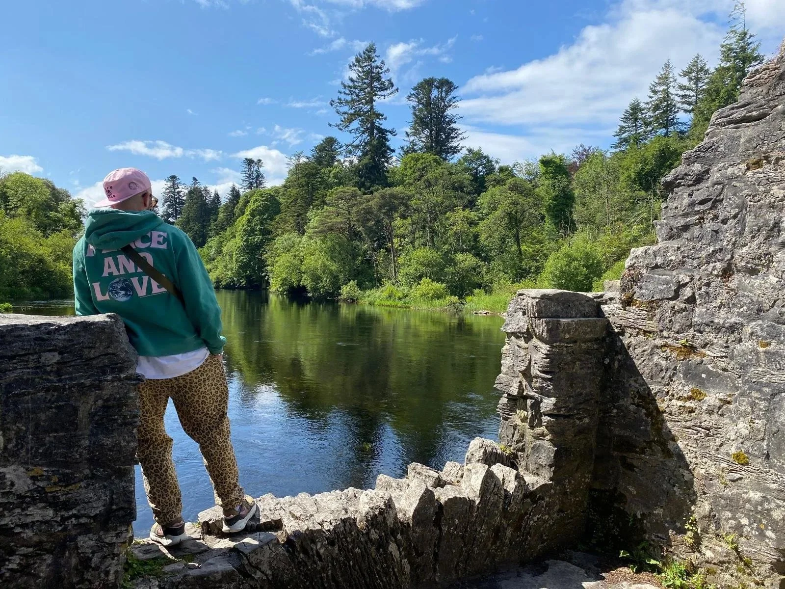 Person standing on a stone bridge by a river, surrounded by lush green trees under a partly cloudy sky. They are wearing Stüssy 'Peace and Love' hoodie, Stüssy 'Pure Space' cap. Complimented with leopard print trousers, Nike Terra vista shoes.