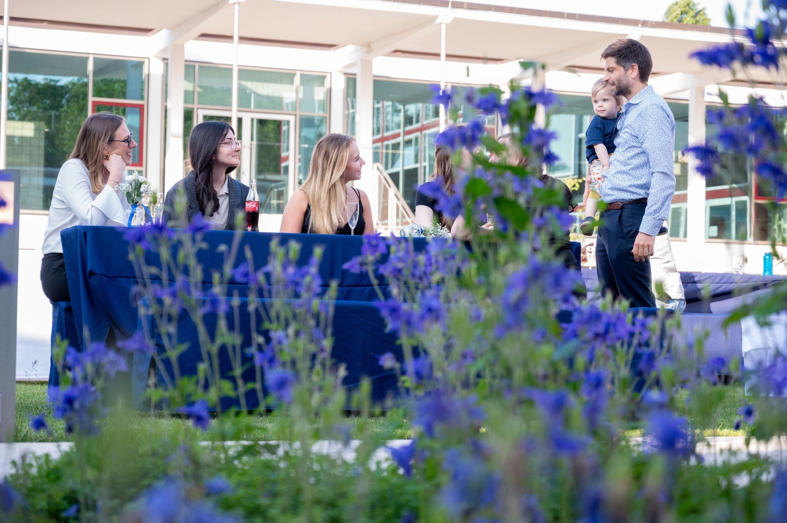 Eine Gruppe von Menschen bei einem Außentreffen, eine Frau steht mit einem Jungen in den Händen, während an einem Tisch sitzende Frauen ihnen zuschauen. Der Bereich ist mit blühenden Blumen im Vordergrund geschmückt.