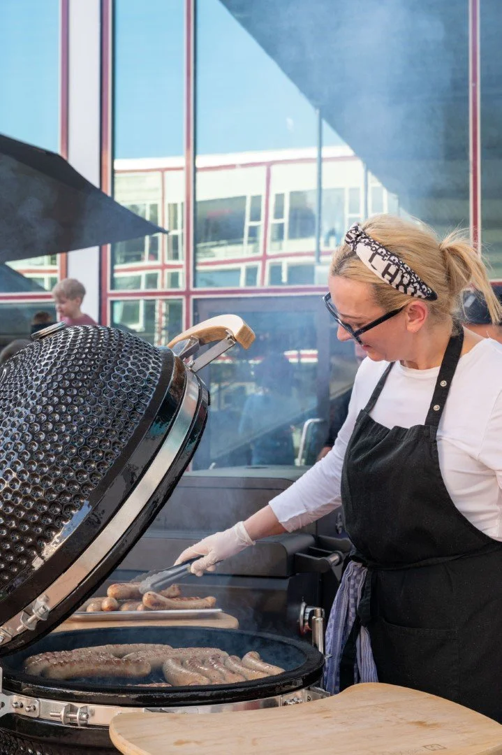 Frau grillt Würstchen auf einem Holzkohlegrill bei einem Außenfest, im Hintergrund ein modernes Gebäude mit großen Fenstern.