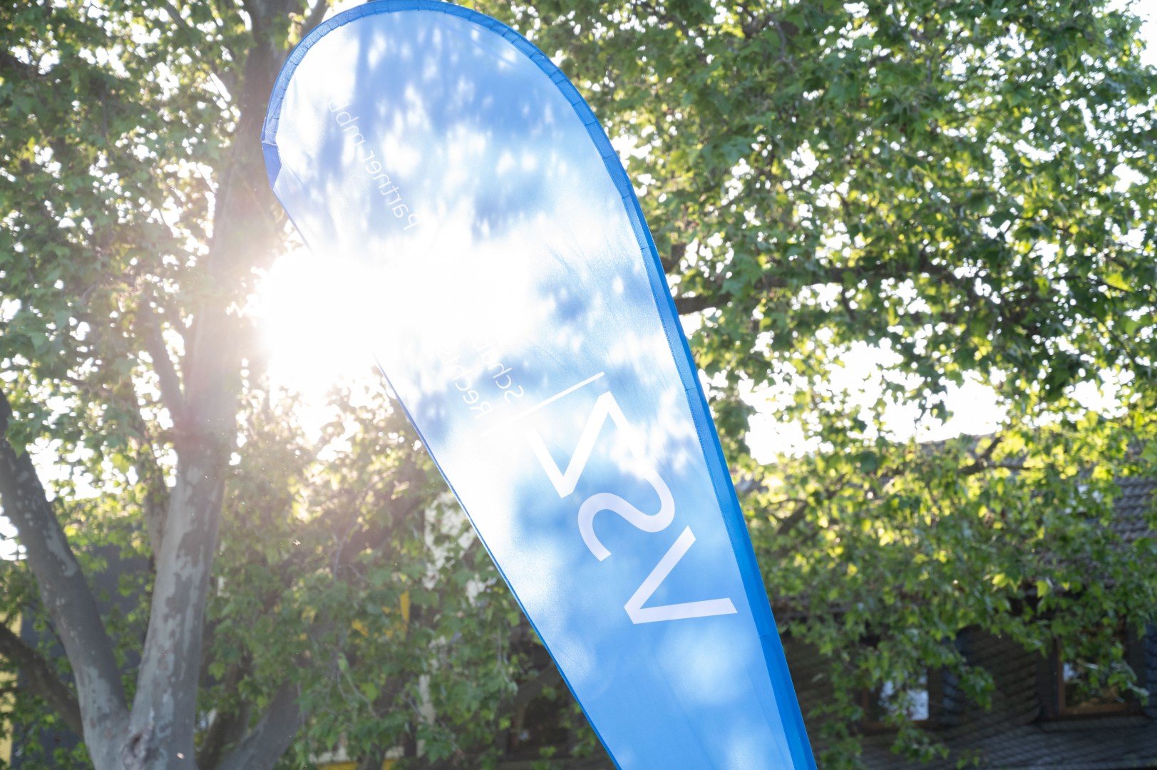 Blauer Fähnchen im Freien, auf dem steht VSZ Rechtsanwälte mit Sonnenlicht und Bäumen im Hintergrund.